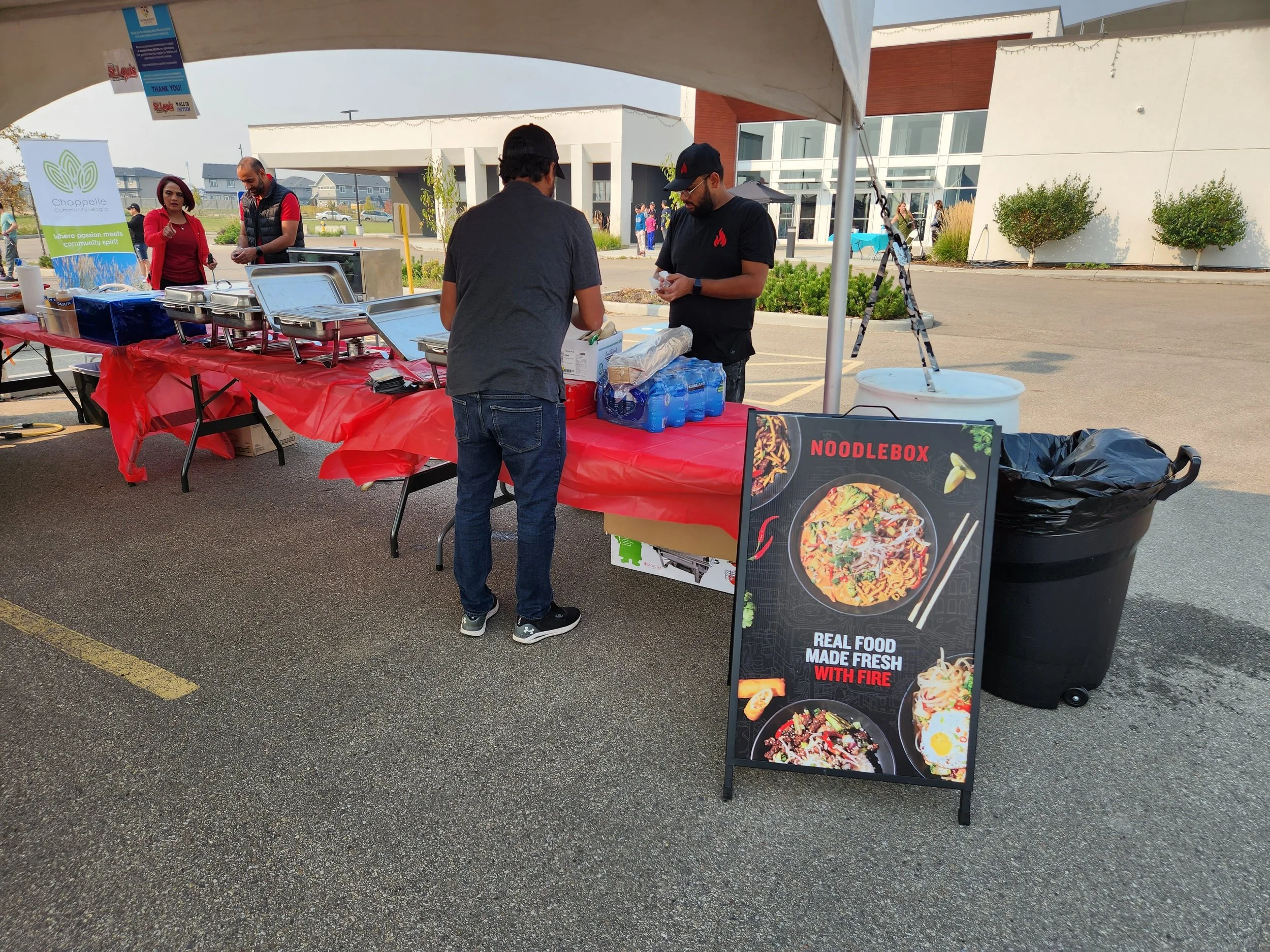 Food stall outdoors under a canopy with a red tablecloth, serving Asian food, with a prominent sign that reads 'NOODLEBOX, REAL FOOD MADE FRESH WITH FIRE'. Two men are approaching the stall, and there are some people in the background.