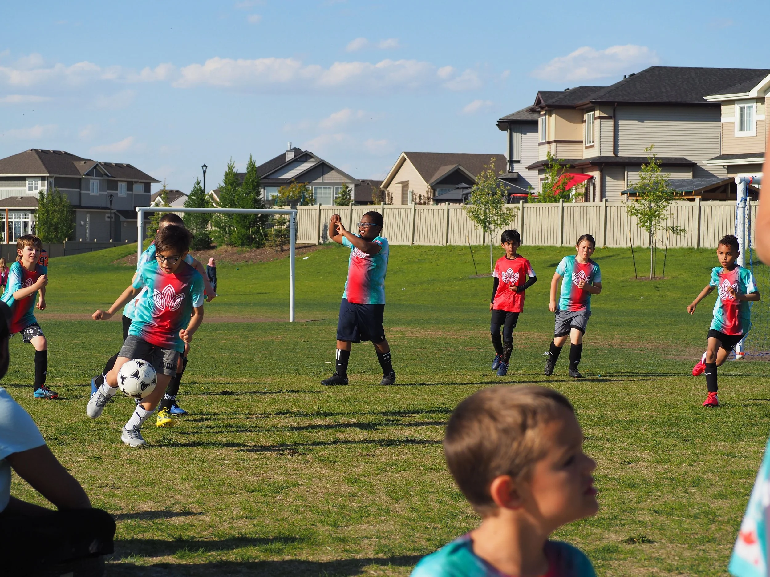 Children playing soccer on a grassy field in a suburban neighborhood, with houses and trees in the background under a blue sky.