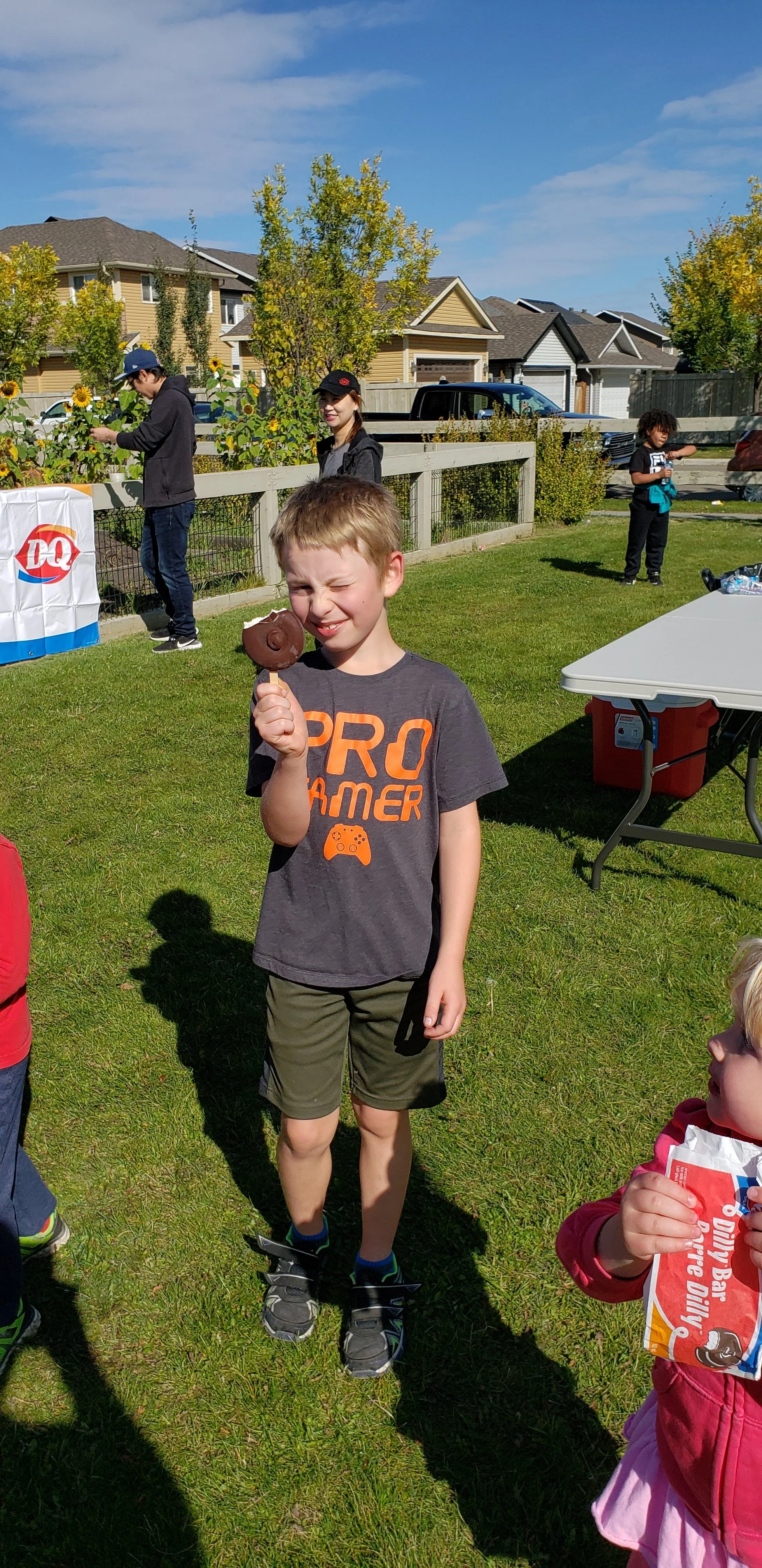 A young boy standing on a grassy area, eating a chocolate-covered ice cream bar, squinting in the sunlight, with other children and houses in the background during an outdoor gathering.