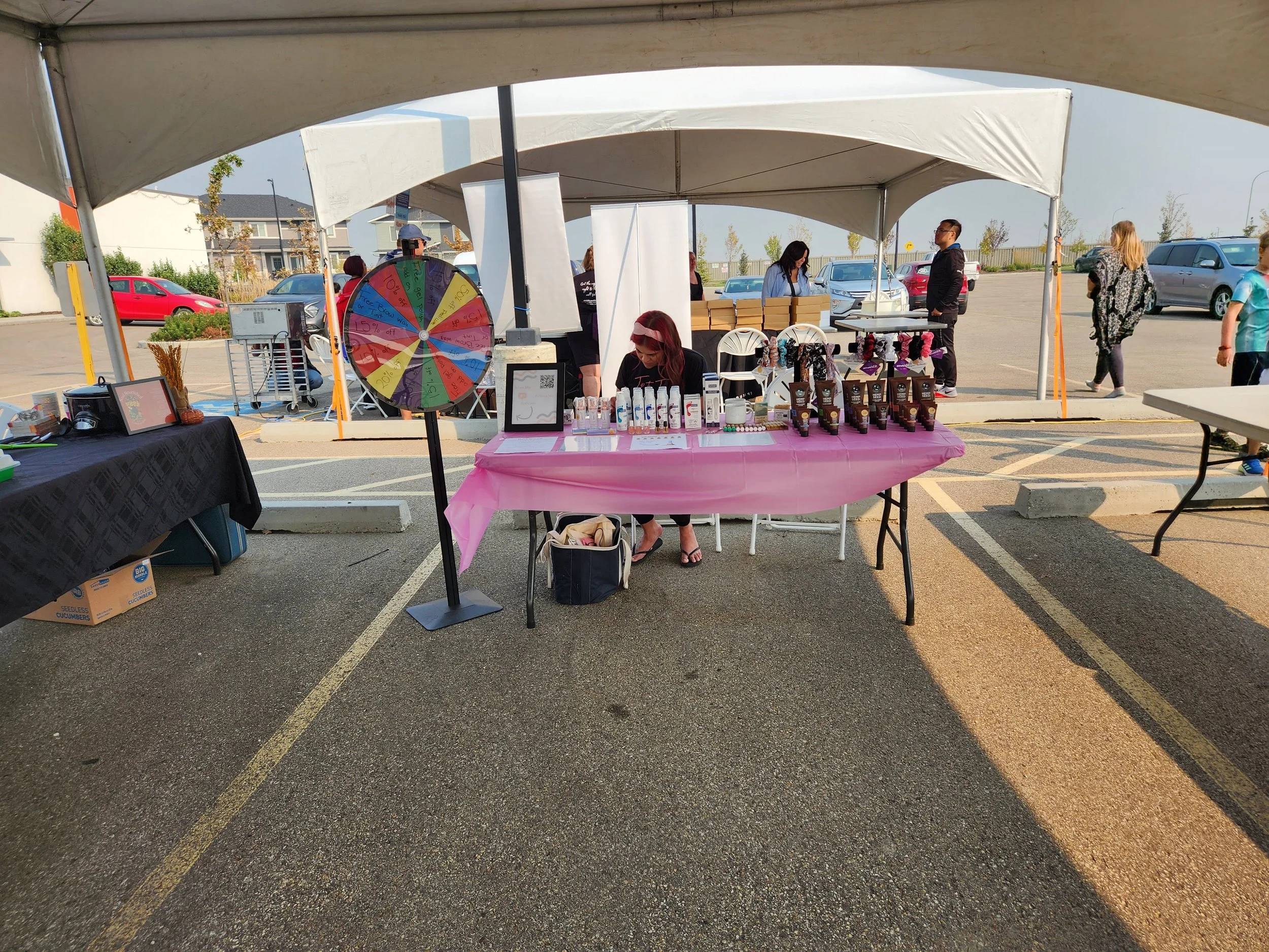 Outdoor market booth with a pink tablecloth displaying various products, a colorful spinning wheel, and a person sitting behind the table. Tents and other people are visible in the background.