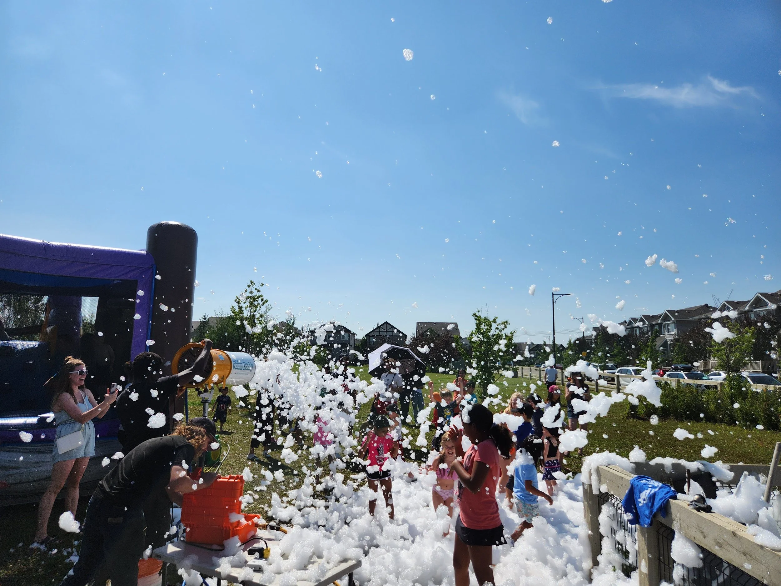 Children playing and laughing in a foam party outdoors on a sunny day with foam, some adults and houses in the background.