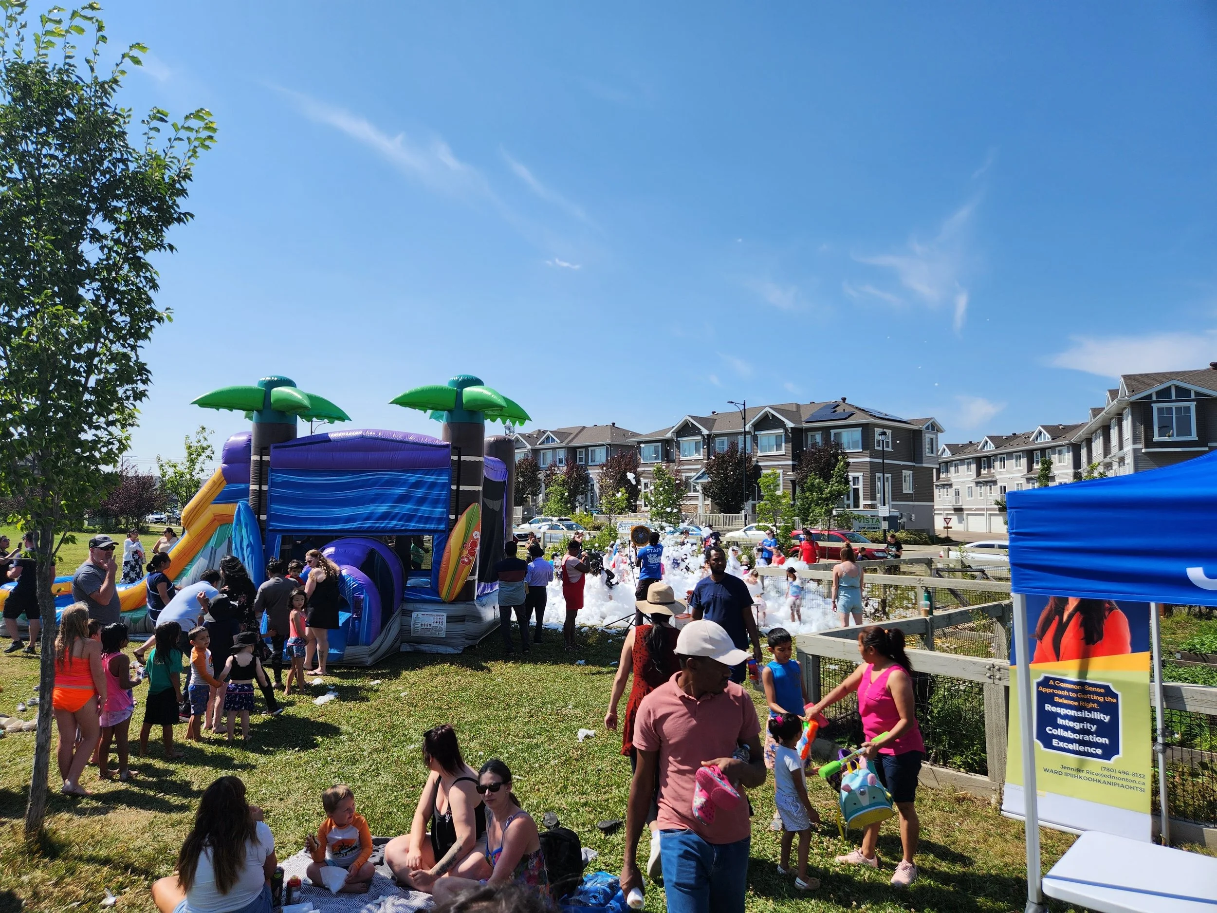 Children and adults at an outdoor summer event with a large inflatable water slide, foam, and picnic activities on a sunny day.