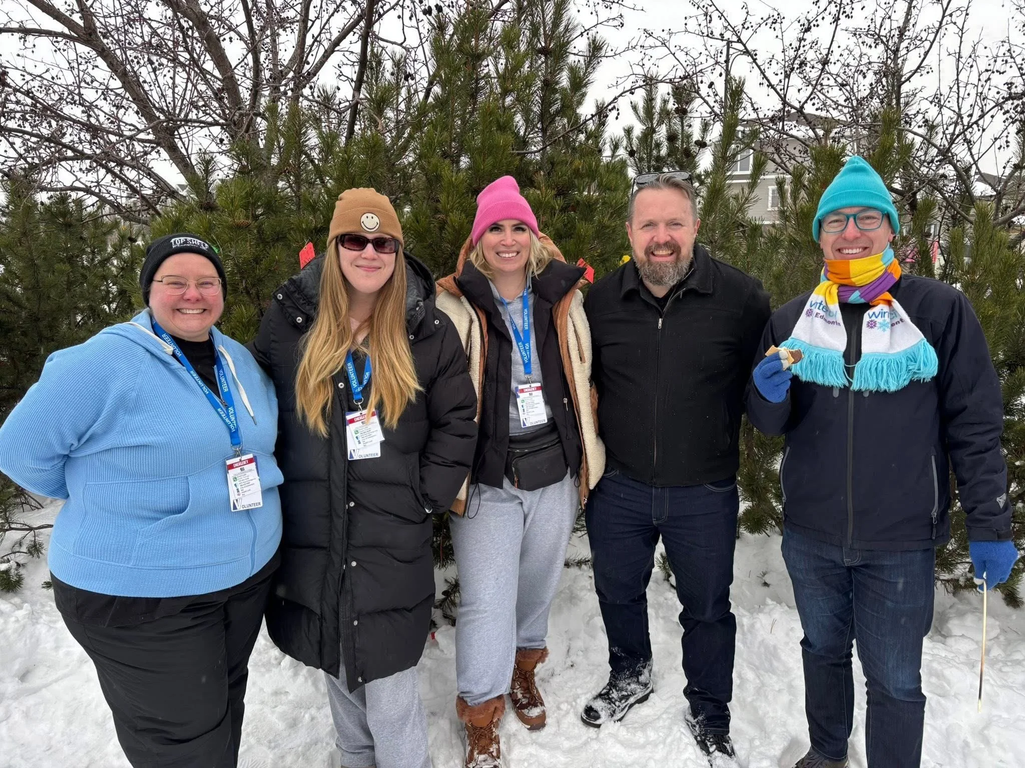 Five people standing outdoors in the snow in front of a pine tree, dressed warmly and smiling at the camera.