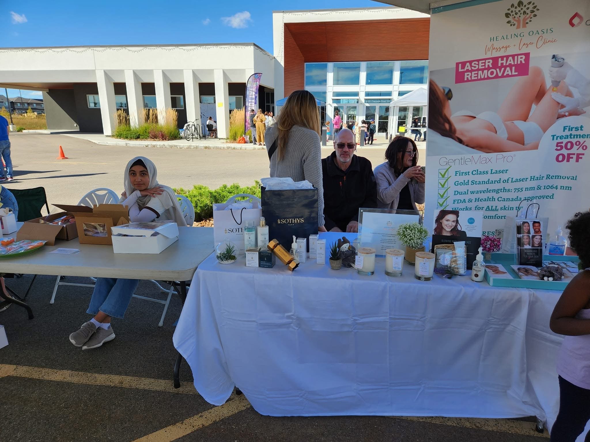 An outdoor health and beauty booth with a large sign advertising laser hair removal, featuring products, promotional materials, and people interacting.