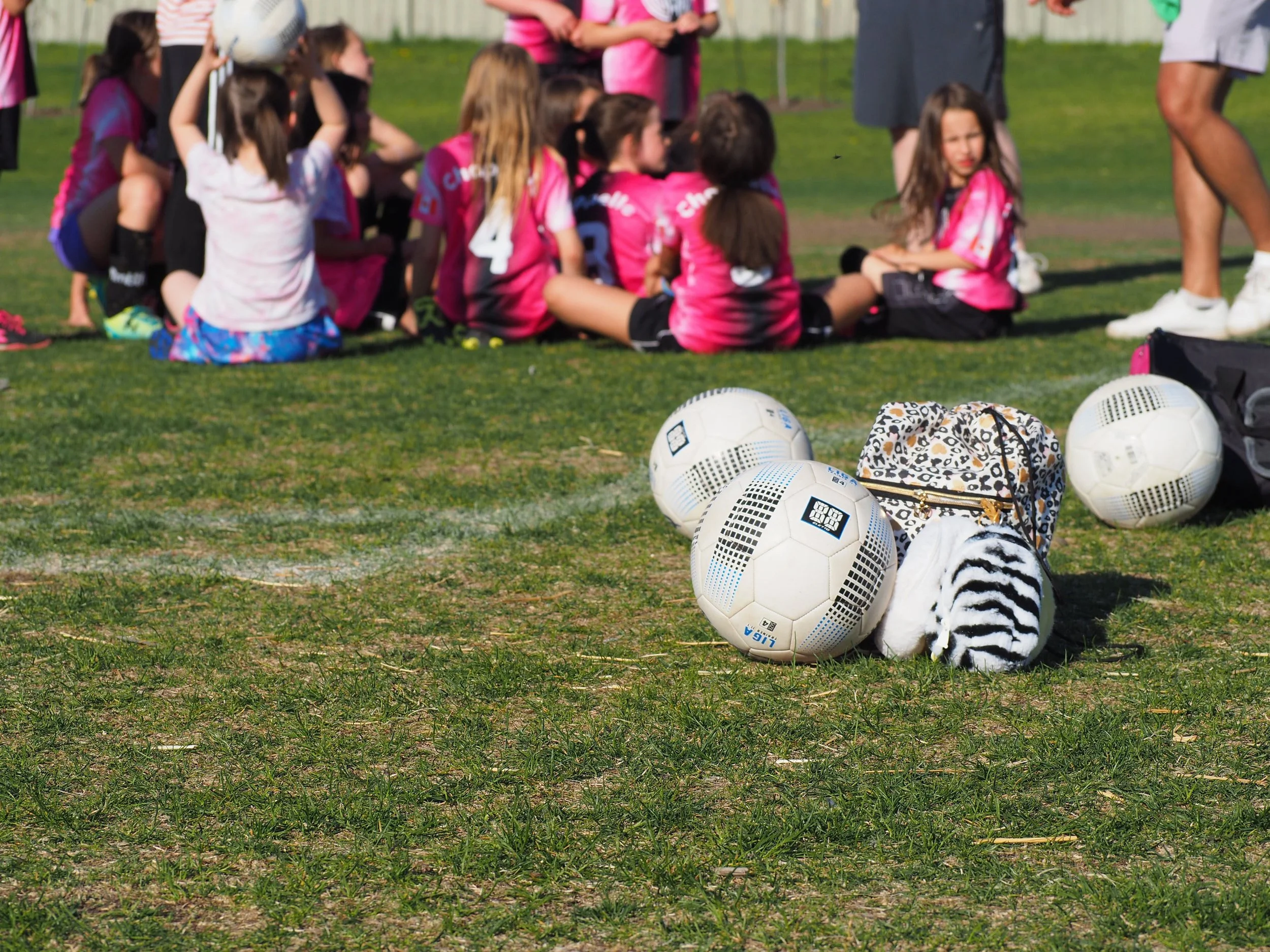 Group of young girls in pink and black sports uniforms sitting on a grassy field after a soccer game, with soccer balls and a leopard-print bag in the foreground.