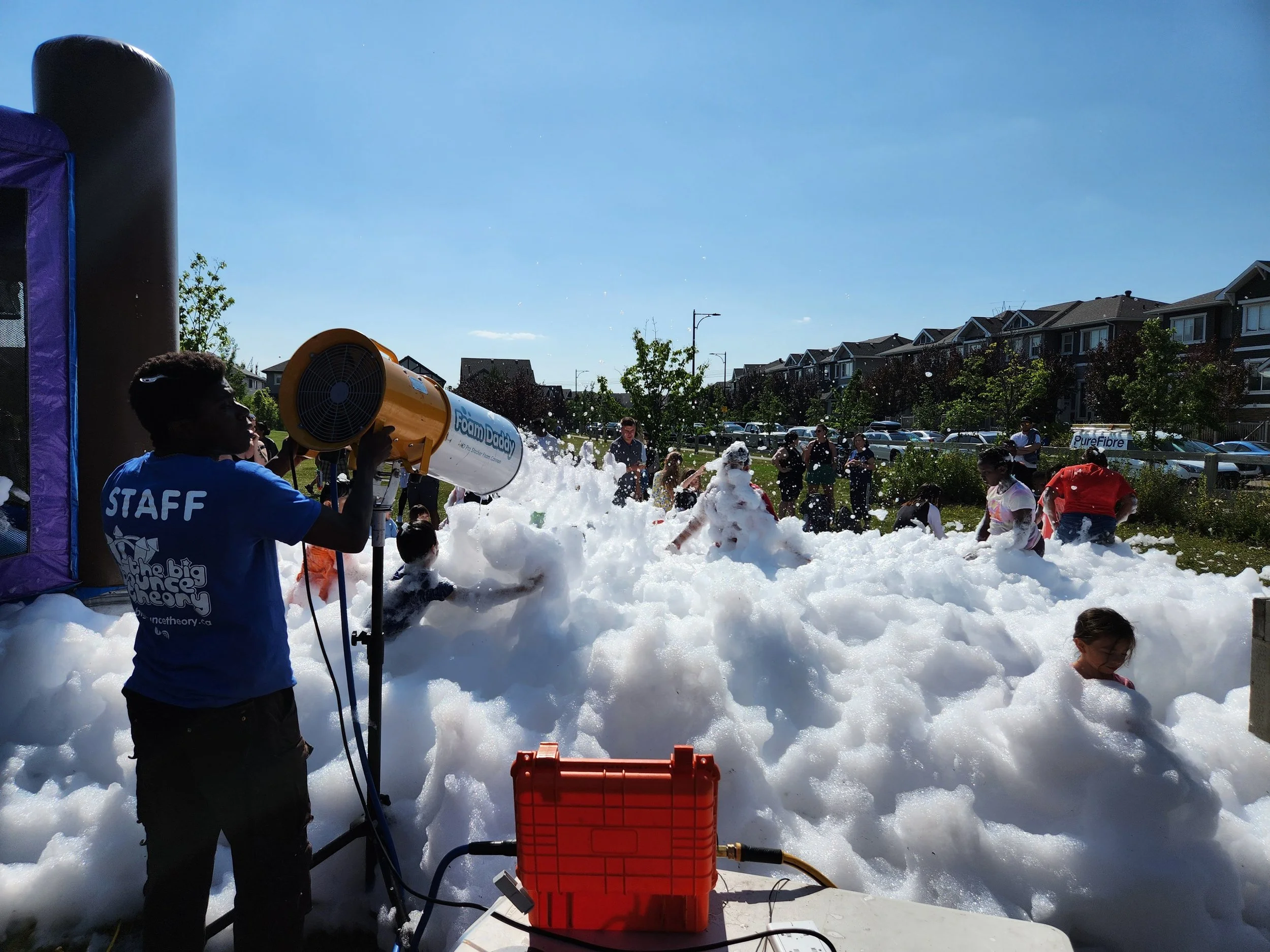 Children and adults playing in a foam pit outdoors, with staff supervising and a foam machine spraying foam, on a sunny day with a row of houses and parked cars in the background.