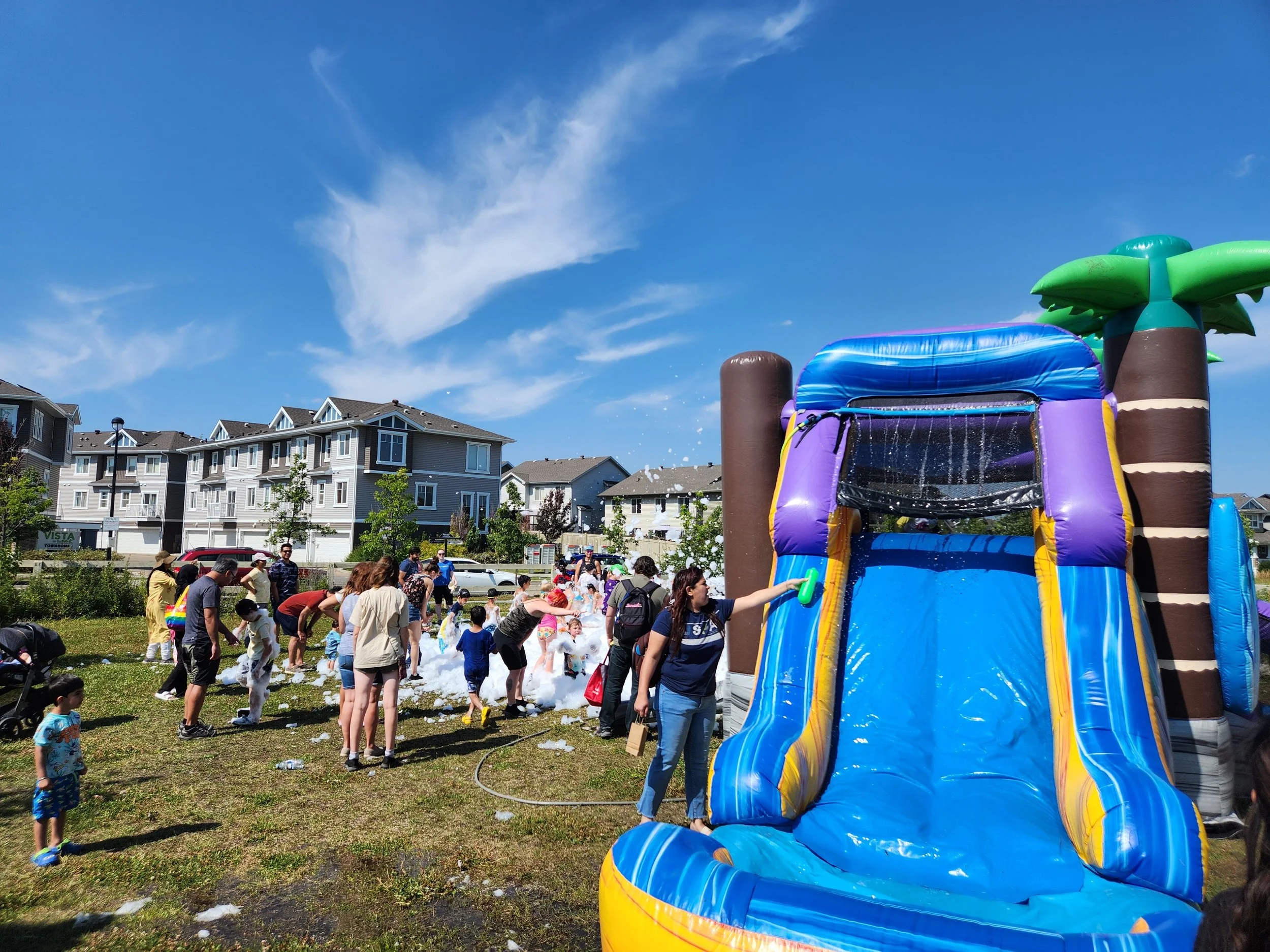 Children and adults playing on a foam bubble pit and inflatable water slide in a grassy park area with residential apartment buildings in the background under a clear blue sky.