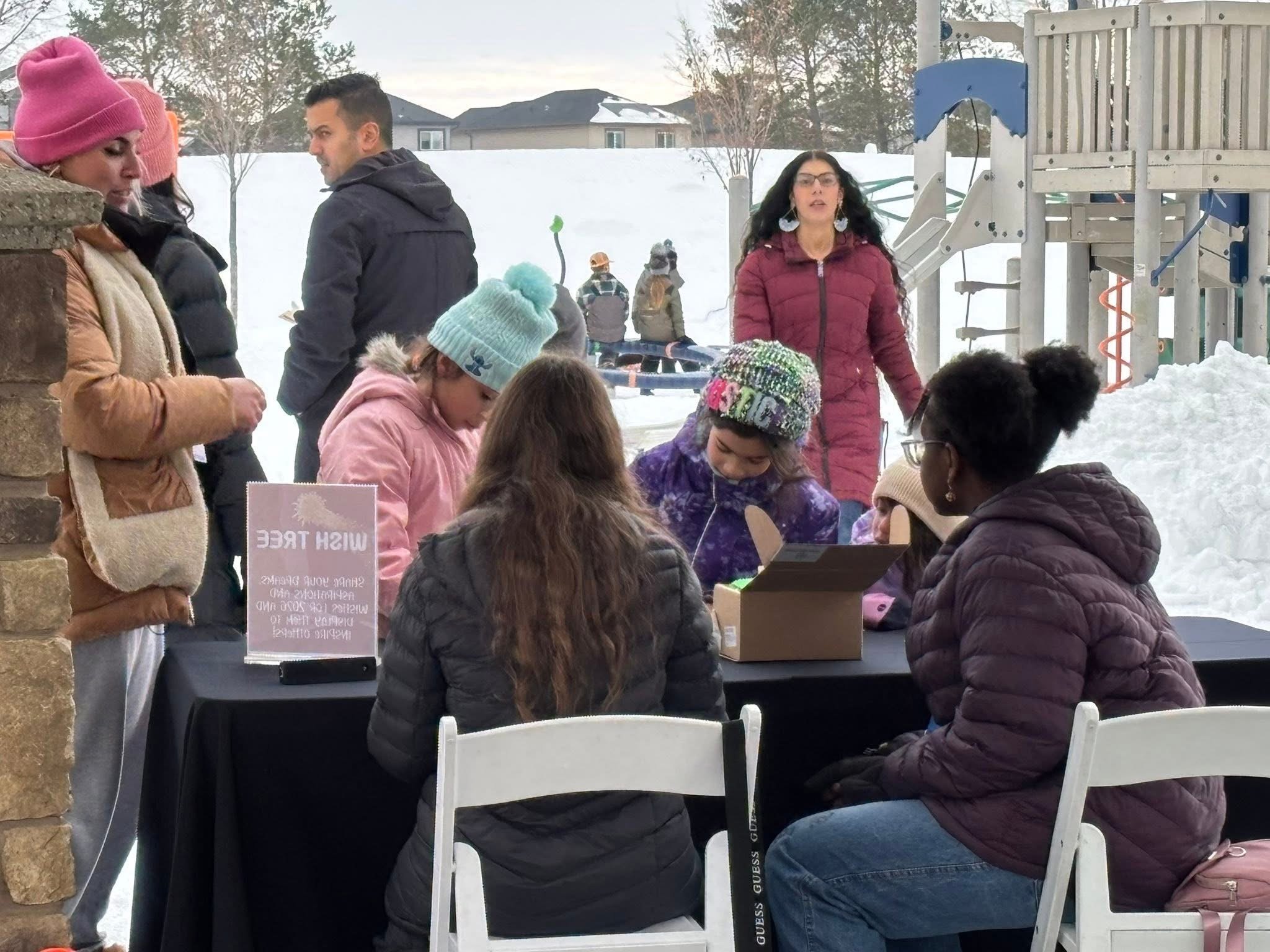 Group of people, including children and adults, gathered around a table for a winter outdoor event, with snow and a playground in the background.