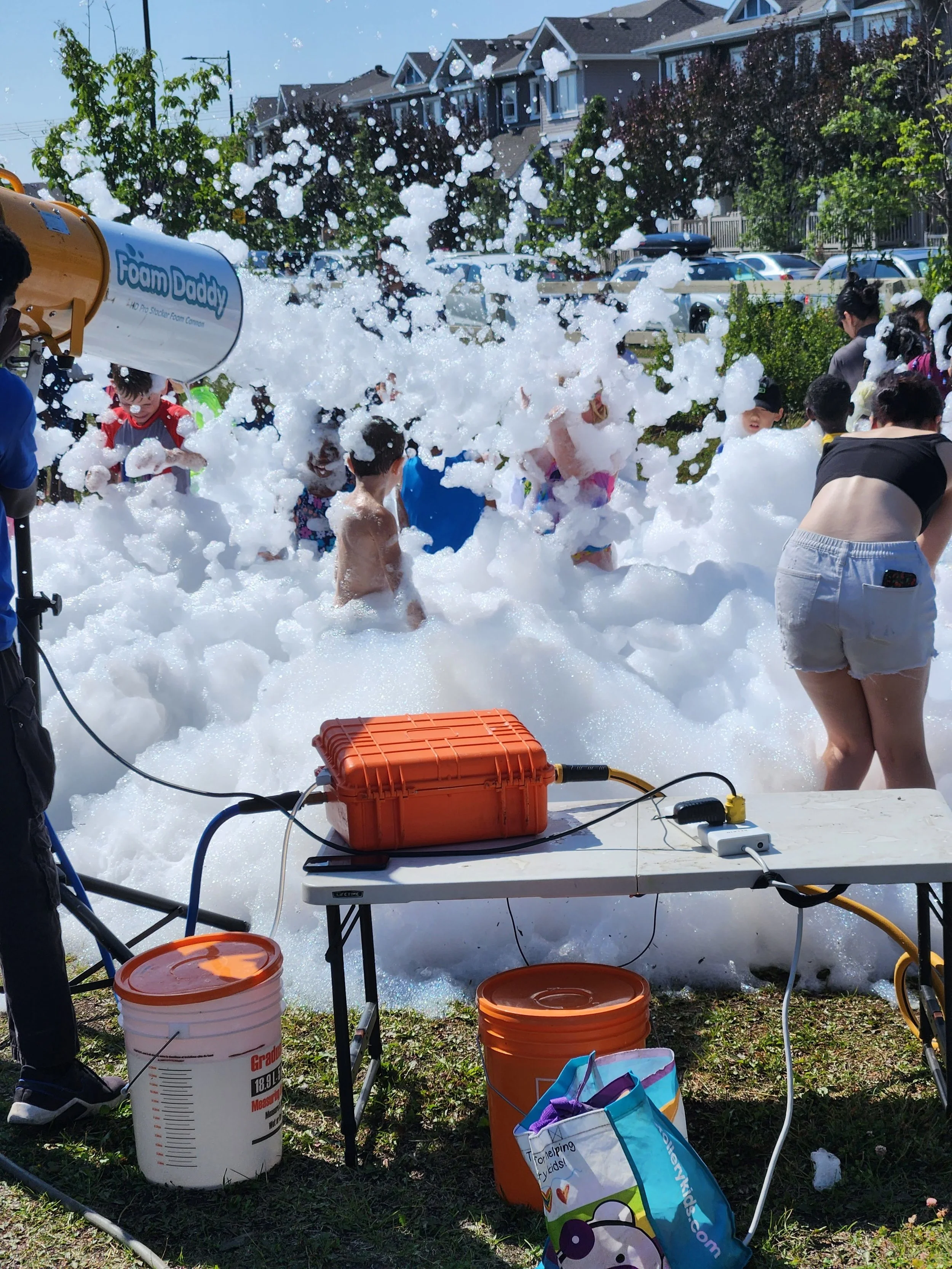 People, including children, playing in foam created by foam machine at outdoor event on a sunny day.