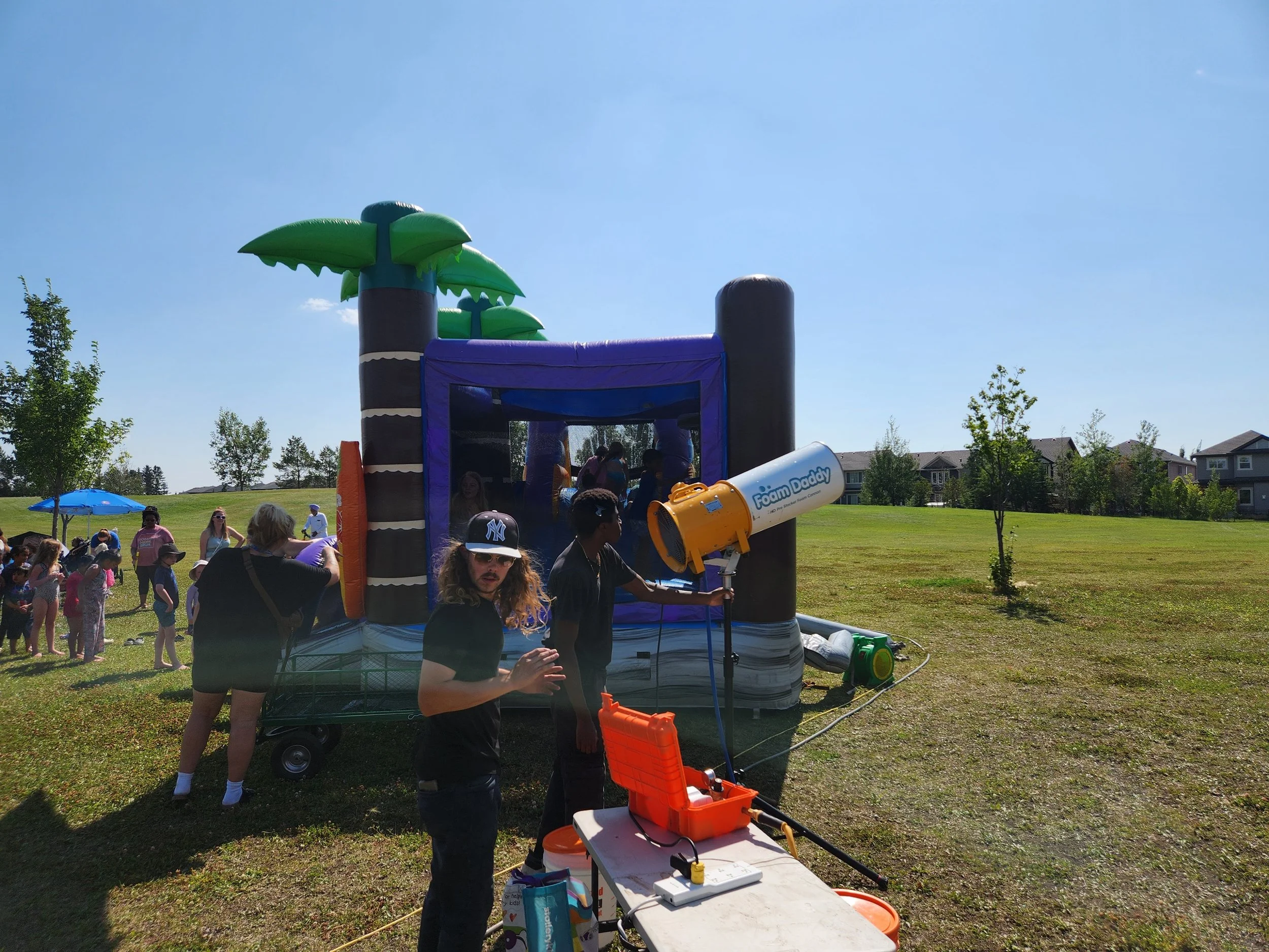 Children and adults gather around a bounce house with palm tree decorations in a grassy park on a sunny day.