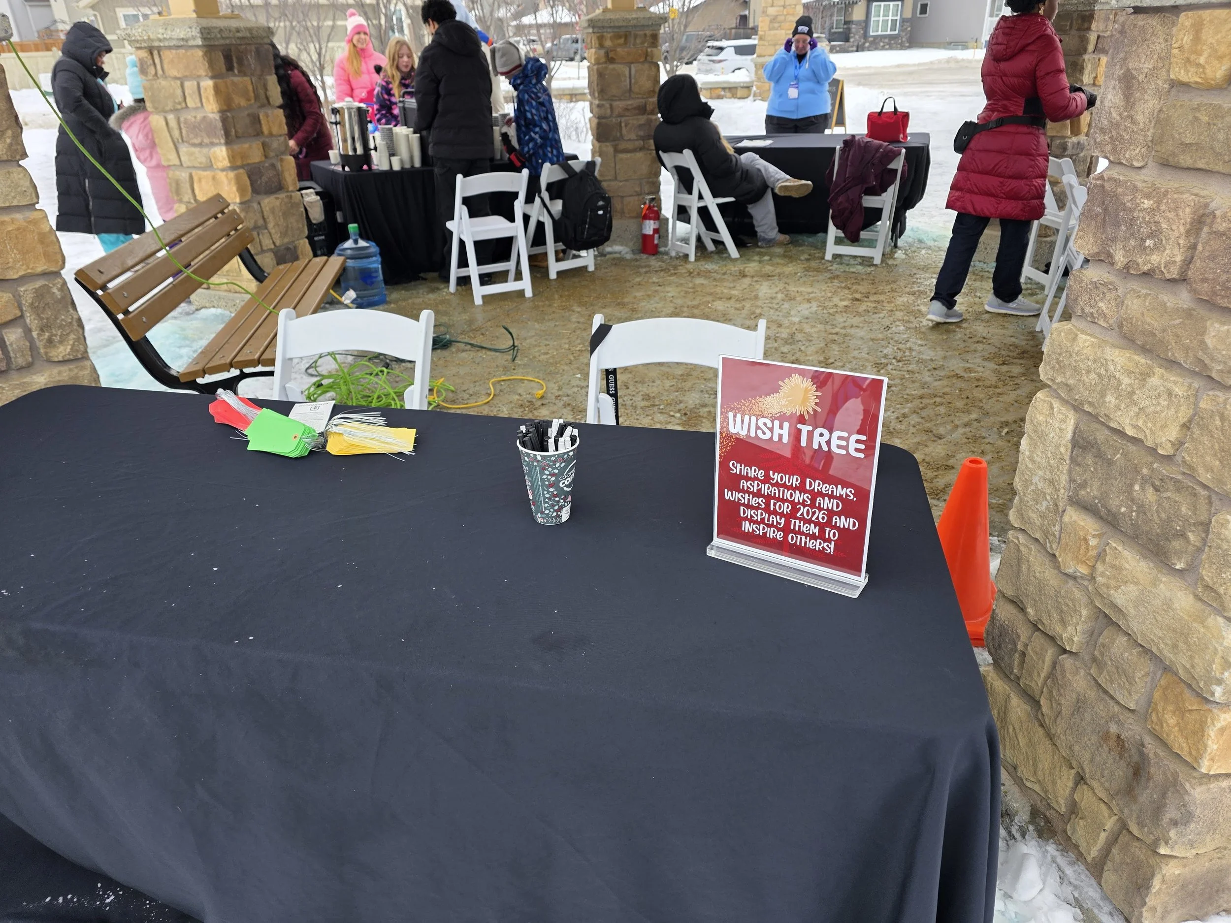 A table covered with a black cloth displaying a sign for a wish tree, with markers and tags. Several people in winter clothing gather in the background under a covered outdoor area with stone pillars, near a snow-covered setting.