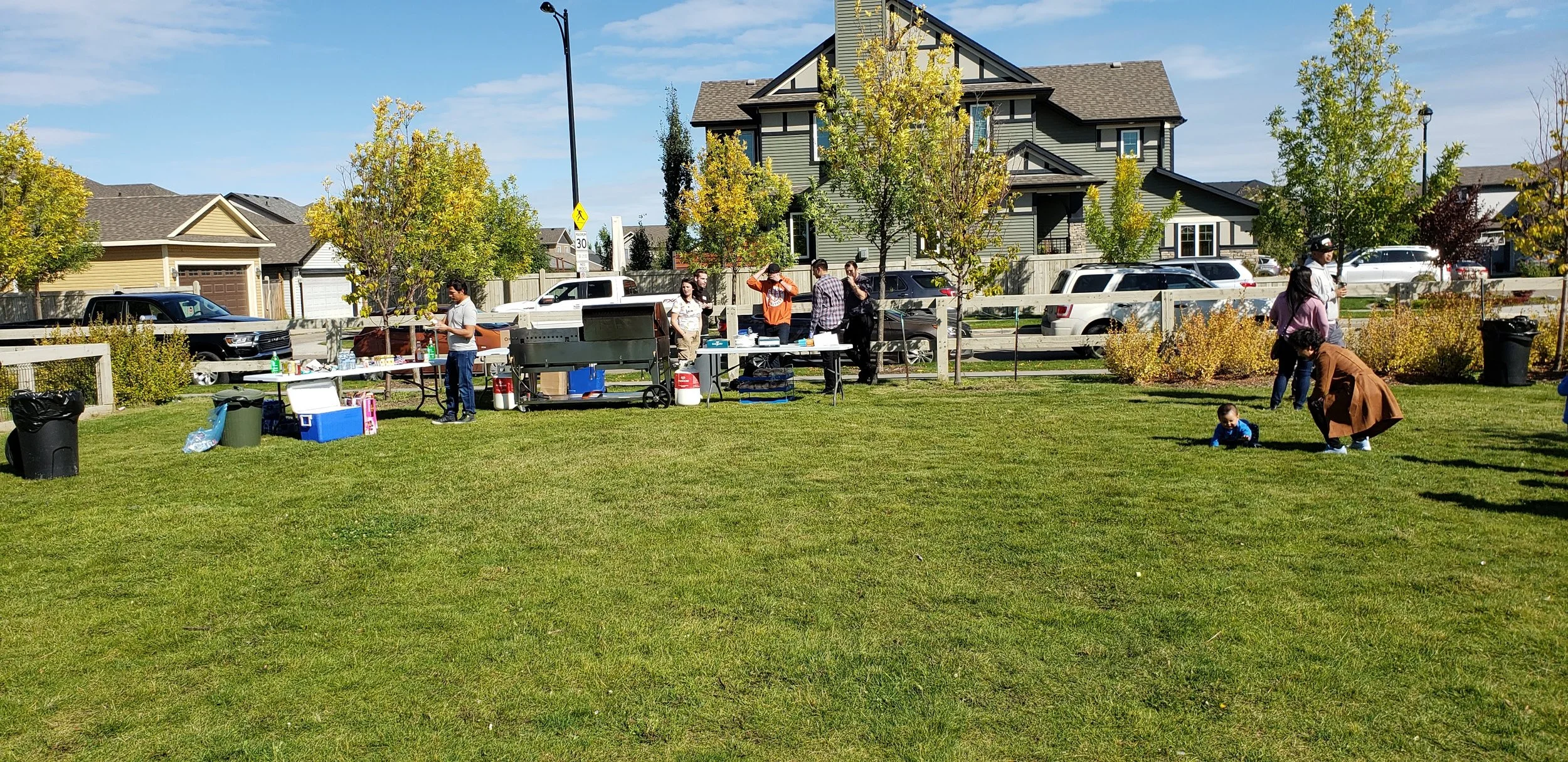 People gathered in a park for a barbecue and picnic with tables, food, and children playing on the grass on a sunny fall day.