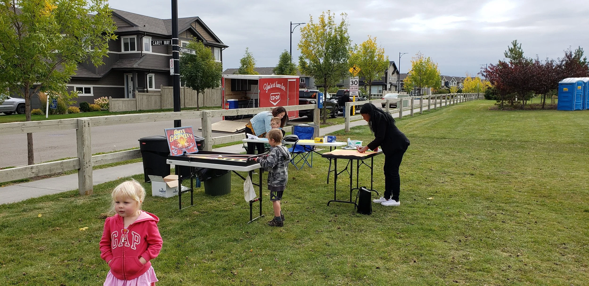 Children and adults setting up or participating in an outdoor event at a park, with tables, game signs, and a grassy area.