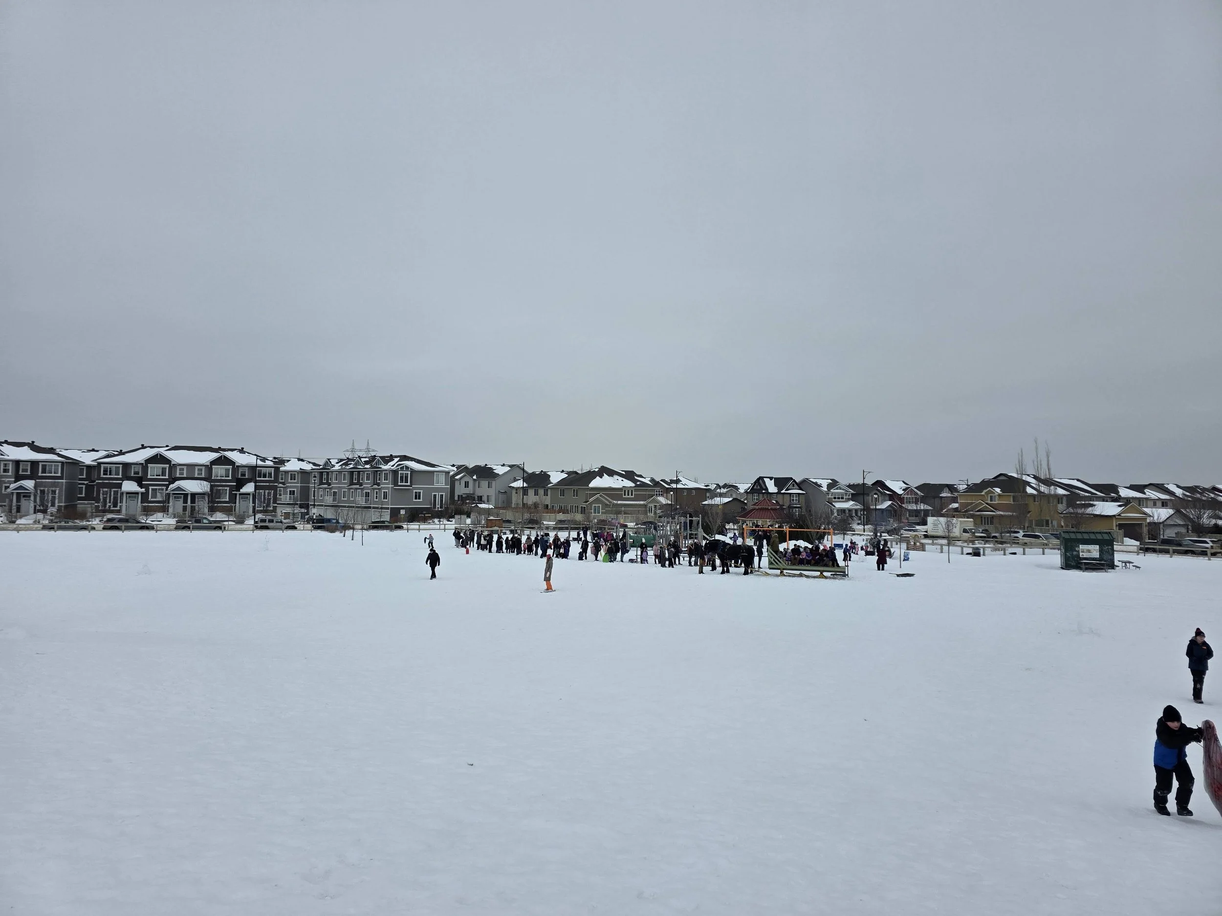 Snow-covered field with people gathered near a playground, winter houses in the background under a grey sky.