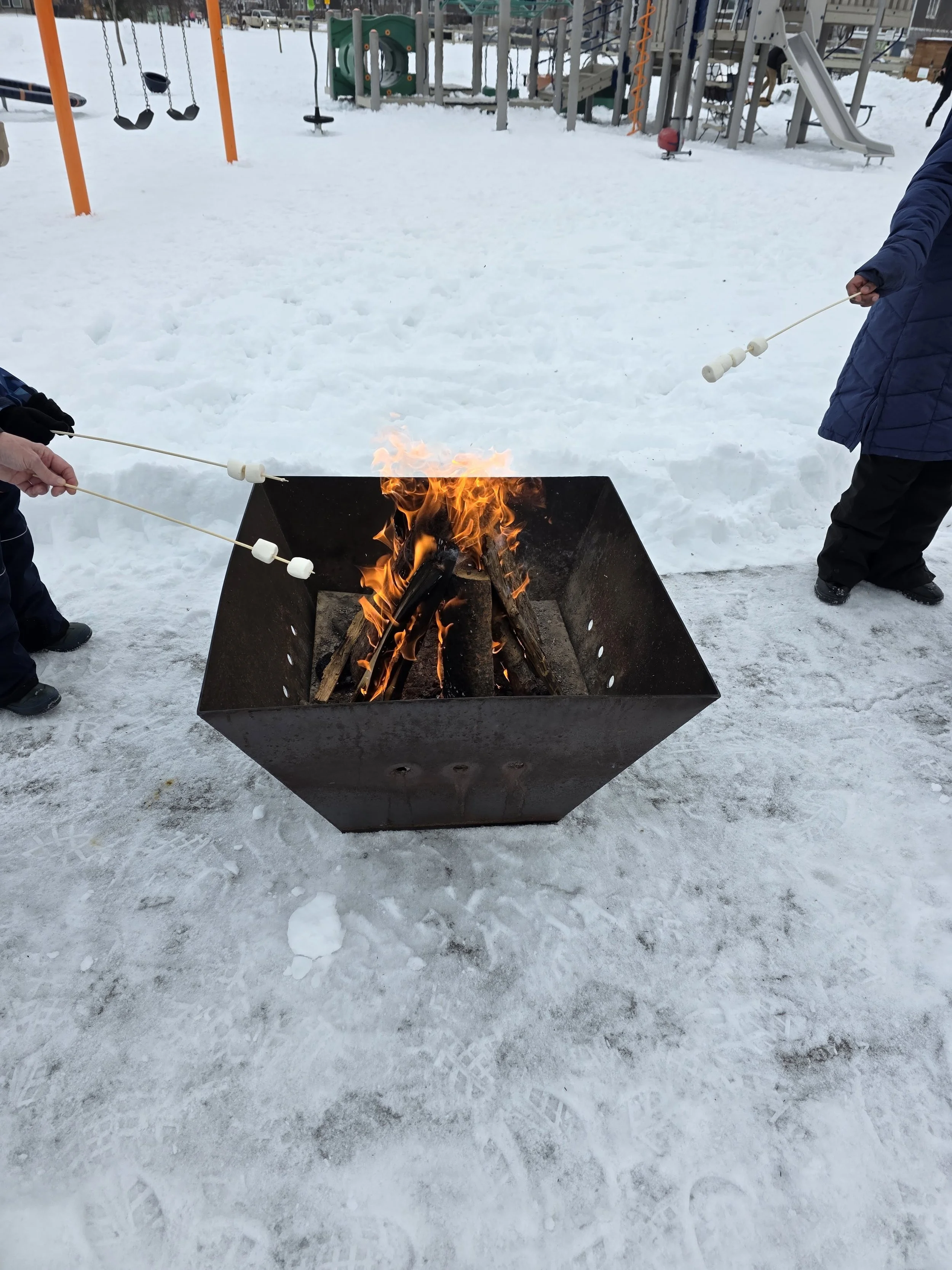 Children roasting marshmallows over an outdoor fire pit in a snowy playground.