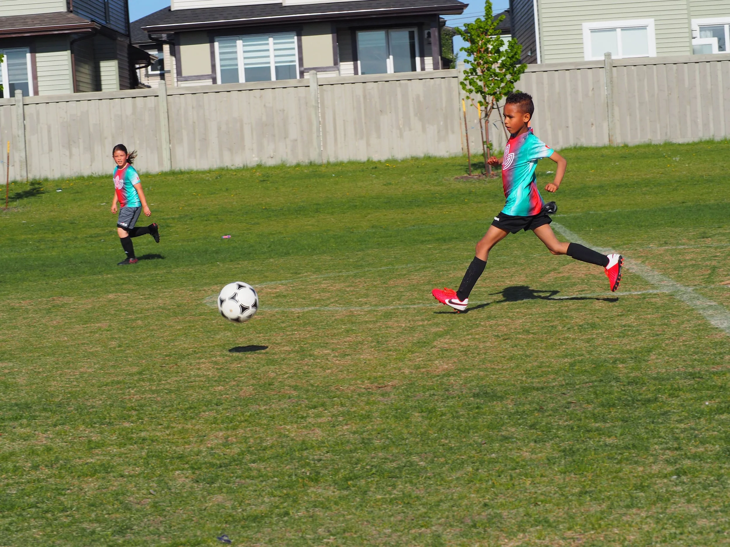 Two children playing soccer on a grassy field near a residential area, with houses and a wooden fence in the background.