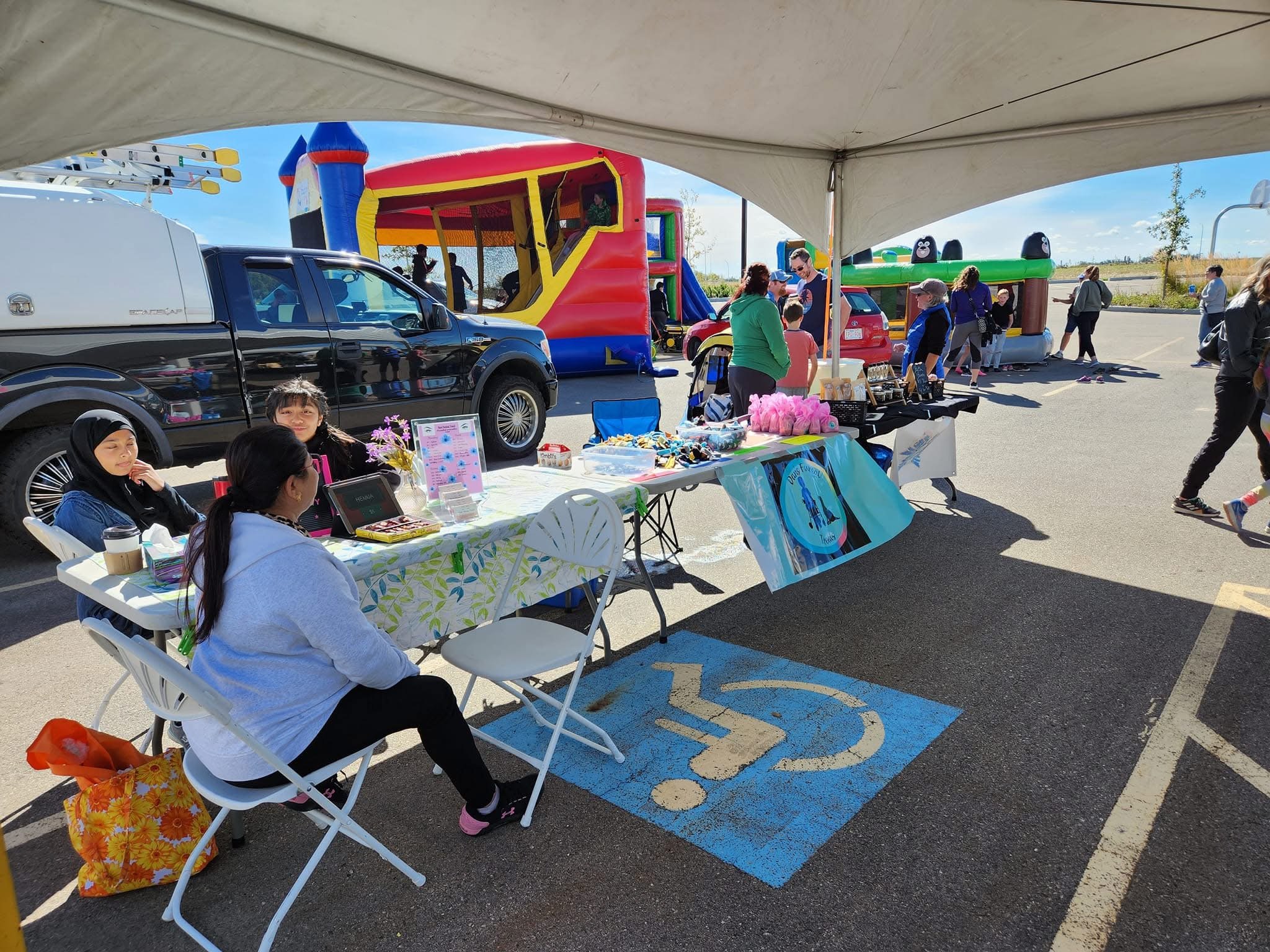 Outdoor market stall set up under a large tent, with tables displaying pink cotton candy, toys, and other items. Several people, including children and adults, are gathered around, some browsing the stall and others enjoying the nearby bouncy houses 