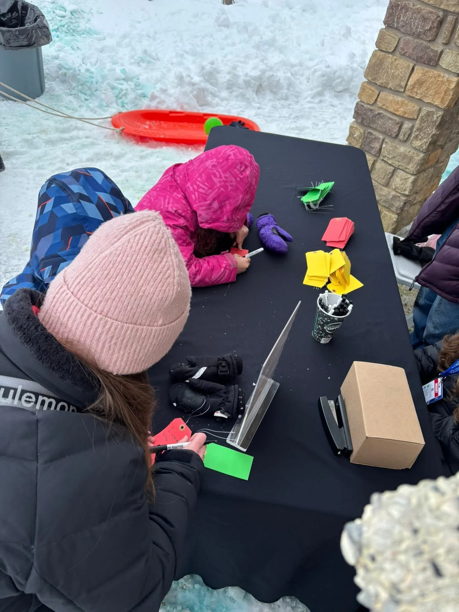 Children playing a game at a table outdoors in winter, with snow on the ground and a sled in the background.