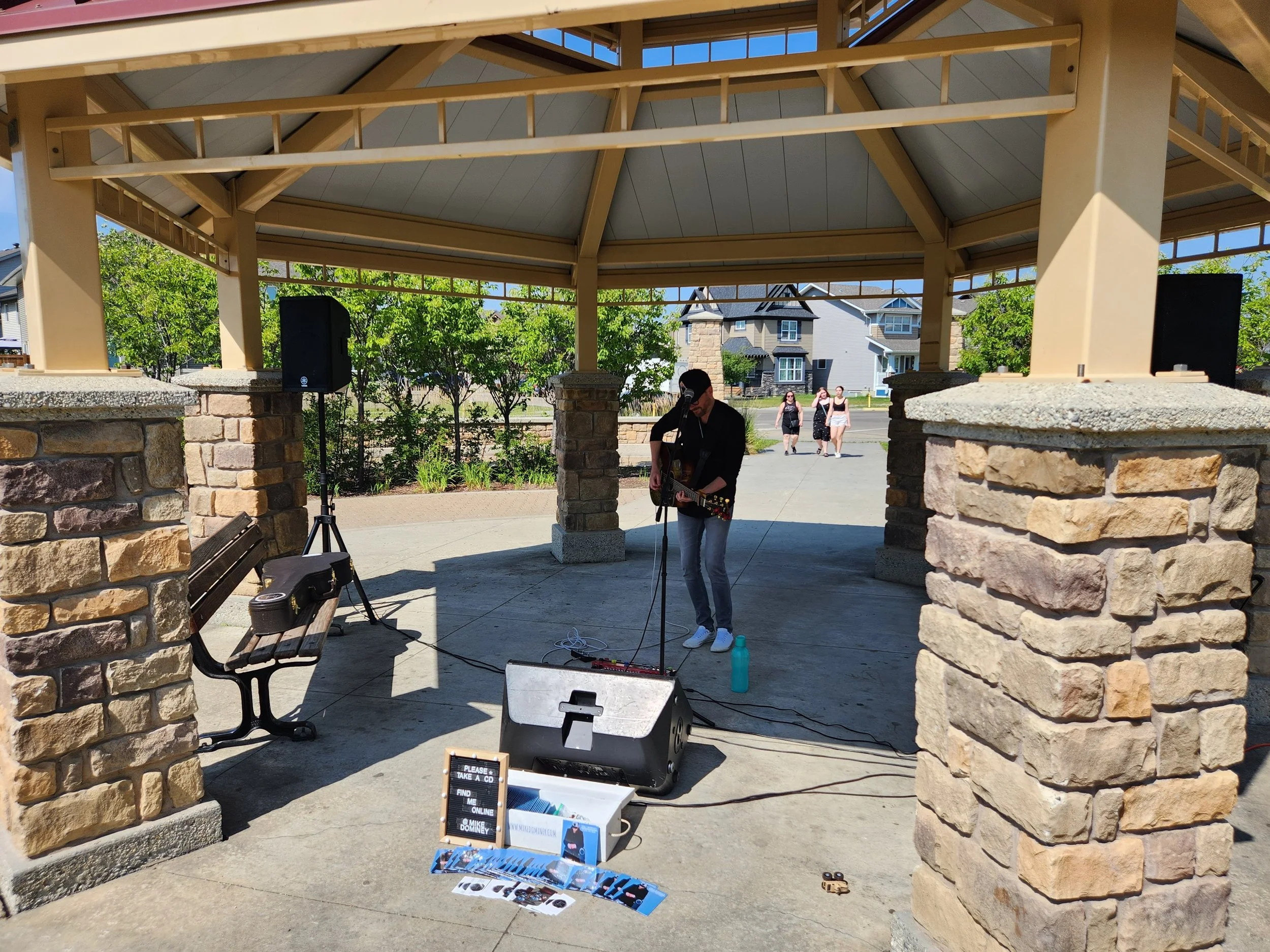 A man playing guitar under a gazebo, with a small crowd walking in the background on a sunny day.