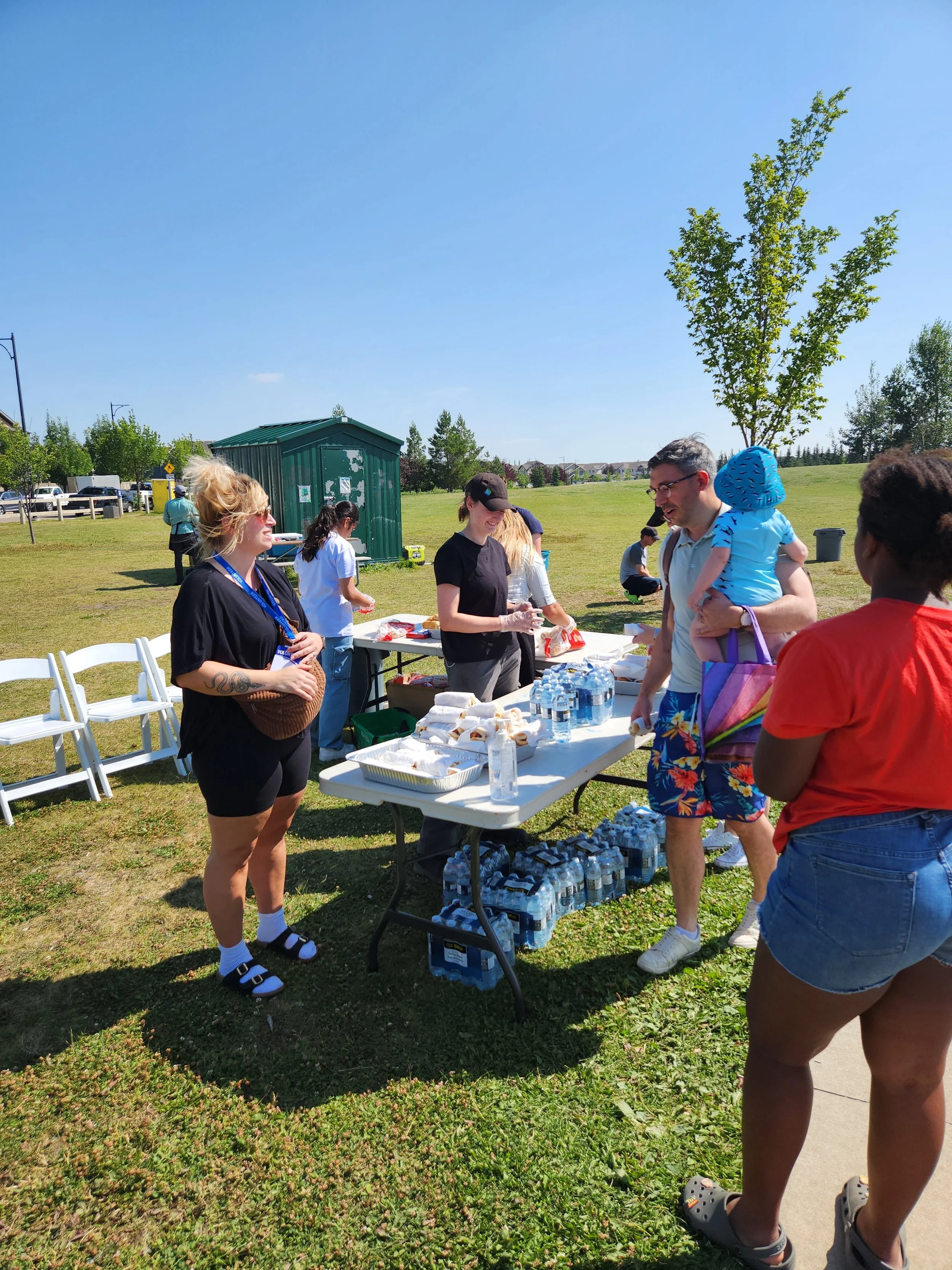 People at an outdoor event with a table of bottled water, snacks, and volunteers handing out items, on a sunny day with green grass and trees.
