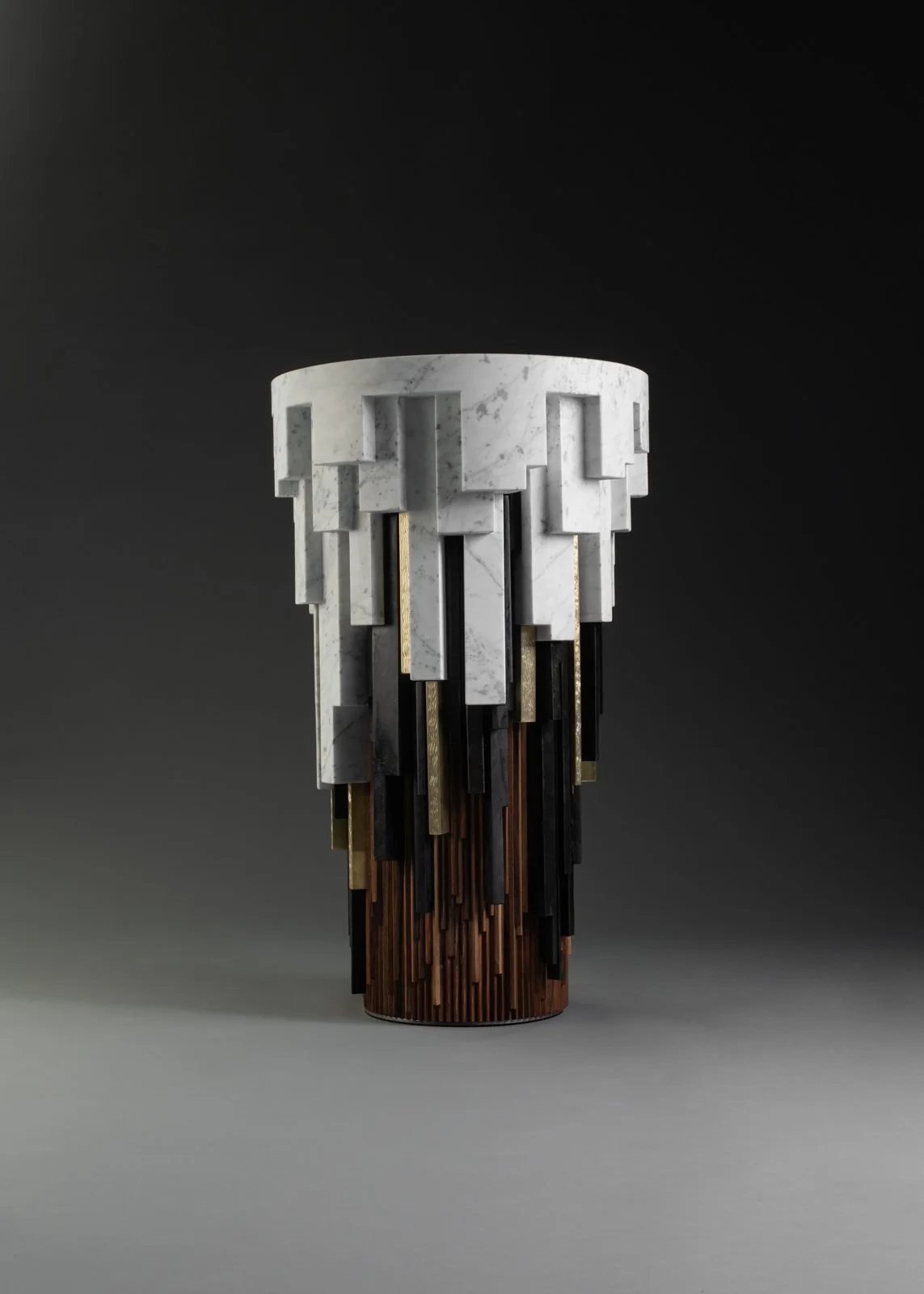 Vase en forme de cylindre avec une partie supérieure en marbre blanc sculptée en forme de motifs géométriques, avec une base composée de segments de bois et de matériaux métalliques en dégradé.