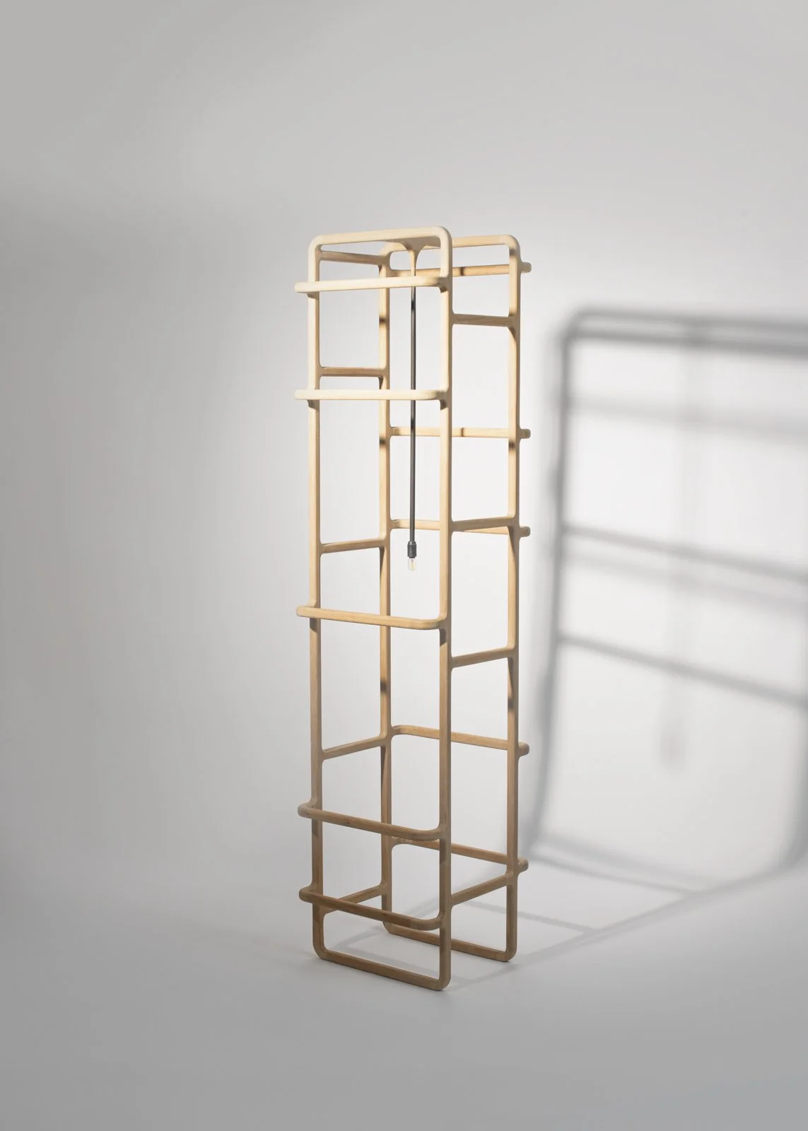 Structure en bois composée de plusieurs cadres rectangulaires empilés, avec un câble électrique suspendu au centre, déployée devant un mur blanc, et une ombre portée à droite.