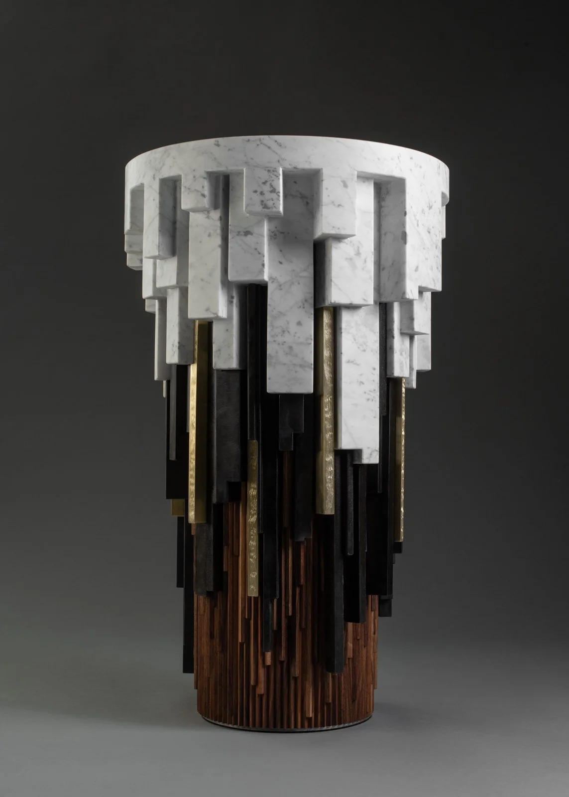 Une sculpture moderne en forme de cylindre composée de blocs de marbre blanc, de bois foncé, et de matériaux métalliques dorés et noirs.