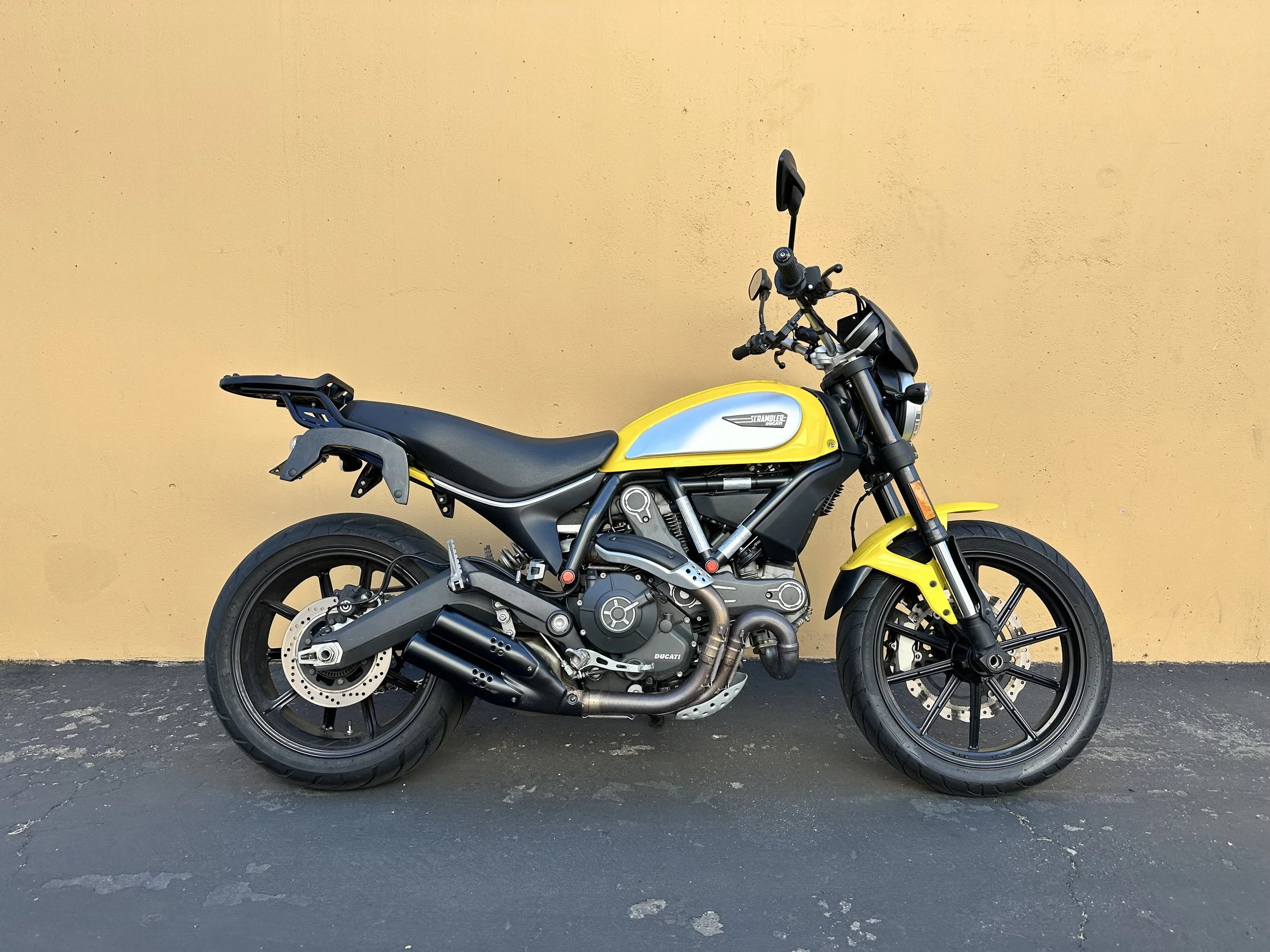 A black and yellow Ducati Scrambler motorcycle parked on asphalt against a beige wall.