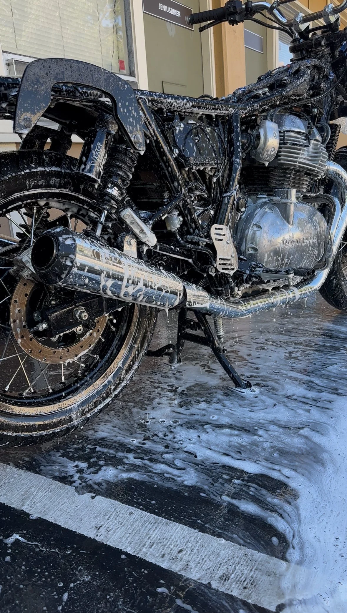 A black motorcycle is being washed with soap and water, with foam and suds on its body, wheels, and exhaust pipe, in a parking lot outside a building.
