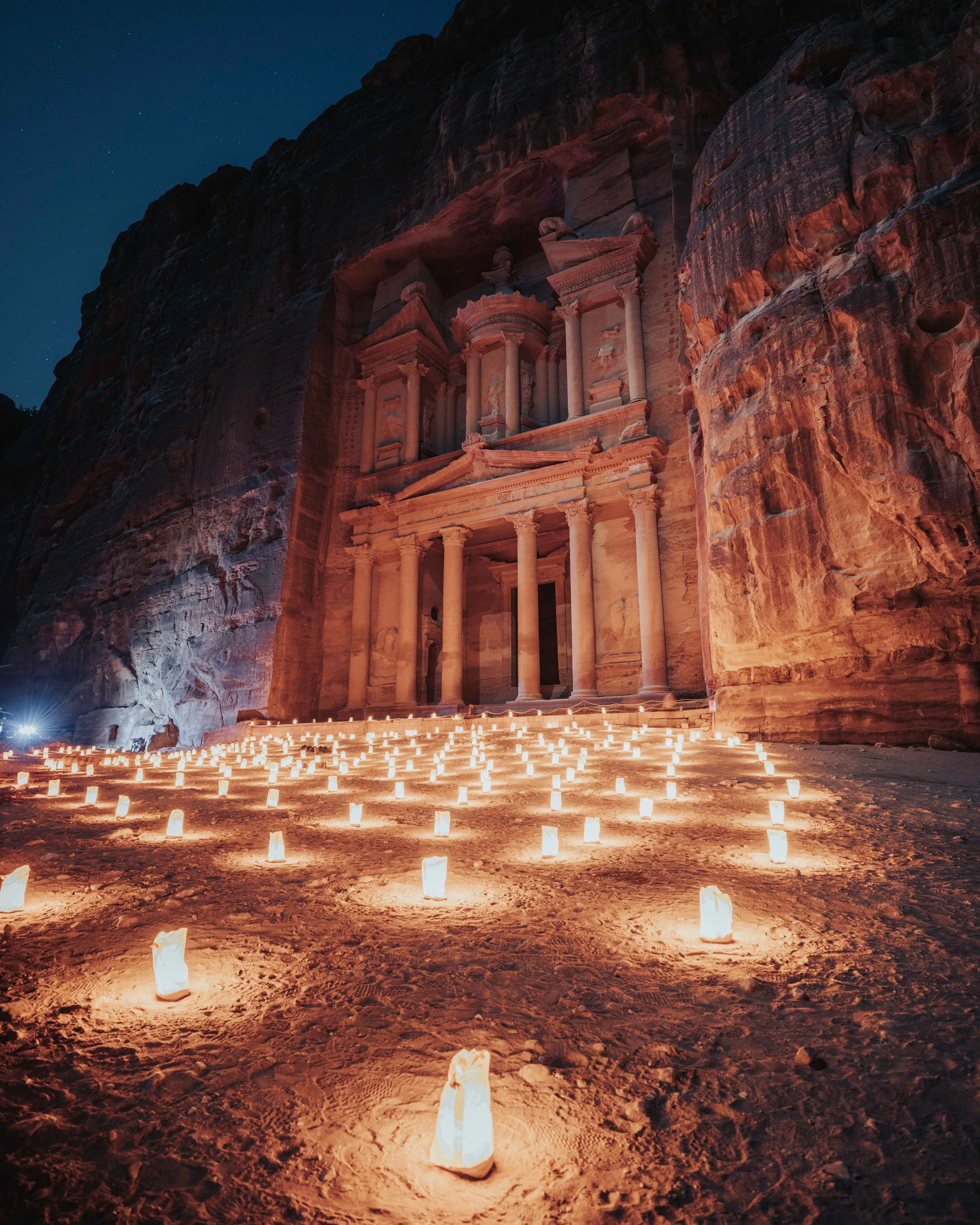 Night view of Petra, Jordan, featuring the iconic rose-red rock-cut architecture illuminated by numerous candles in paper lanterns on the sandy ground.