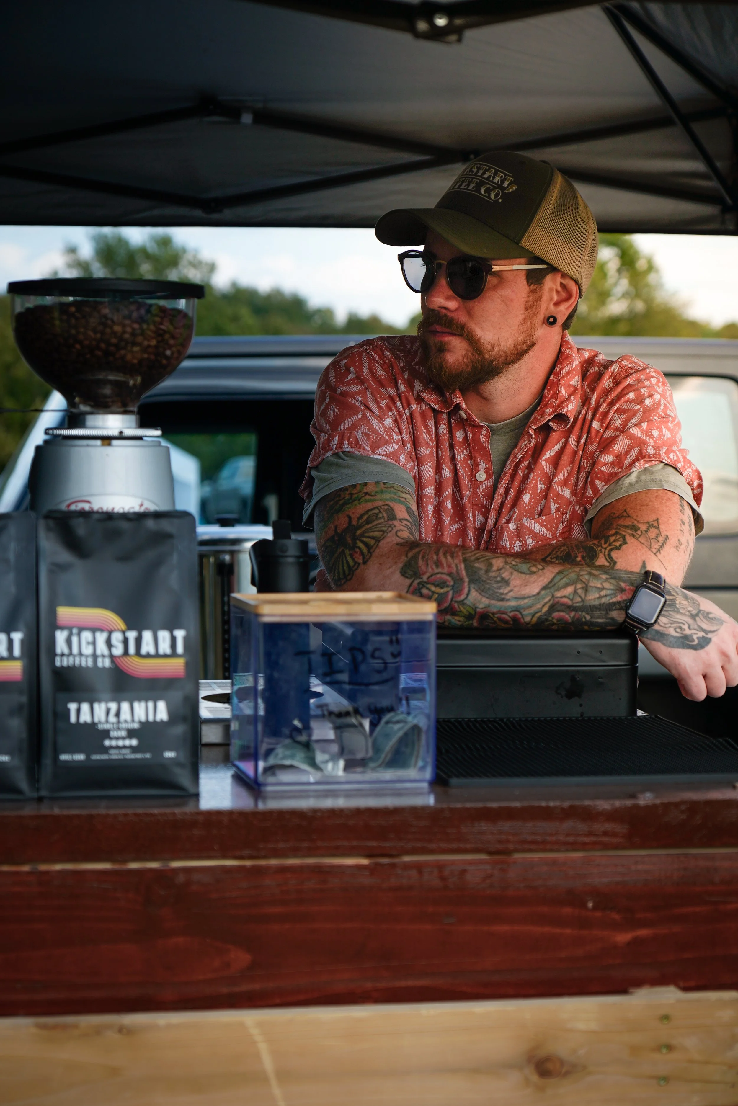 A man with tattoos, wearing sunglasses and a cap, stands behind a coffee stand under a black canopy, with bags of coffee, a coffee grinder, and a cash box on the counter, outdoors with trees in the background.
