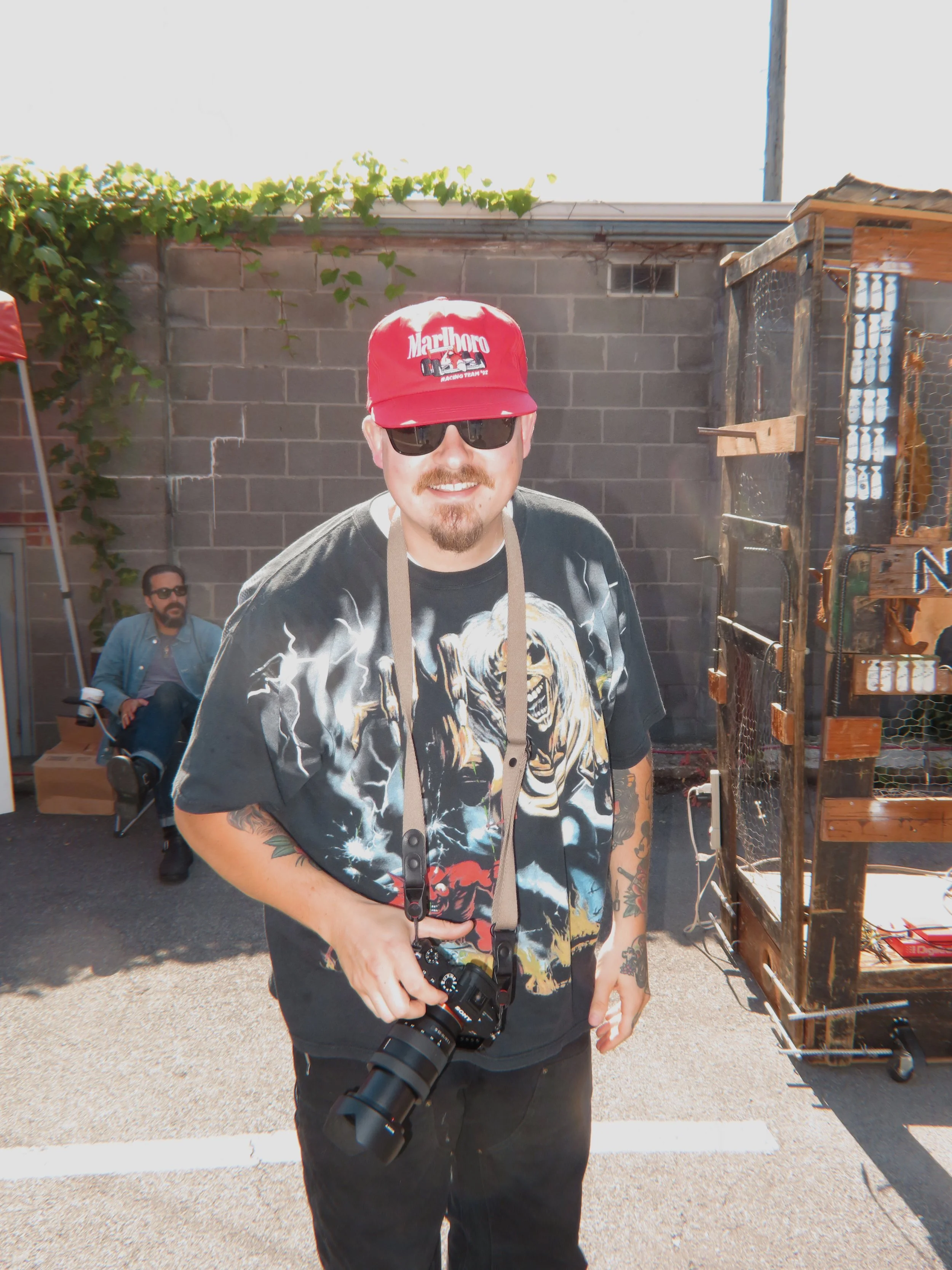 Young man wearing sunglasses, red Marlboro cap, and graphic t-shirt, holding a camera outdoors in a sunny setting. In the background, a seated man wears sunglasses and a denim jacket.