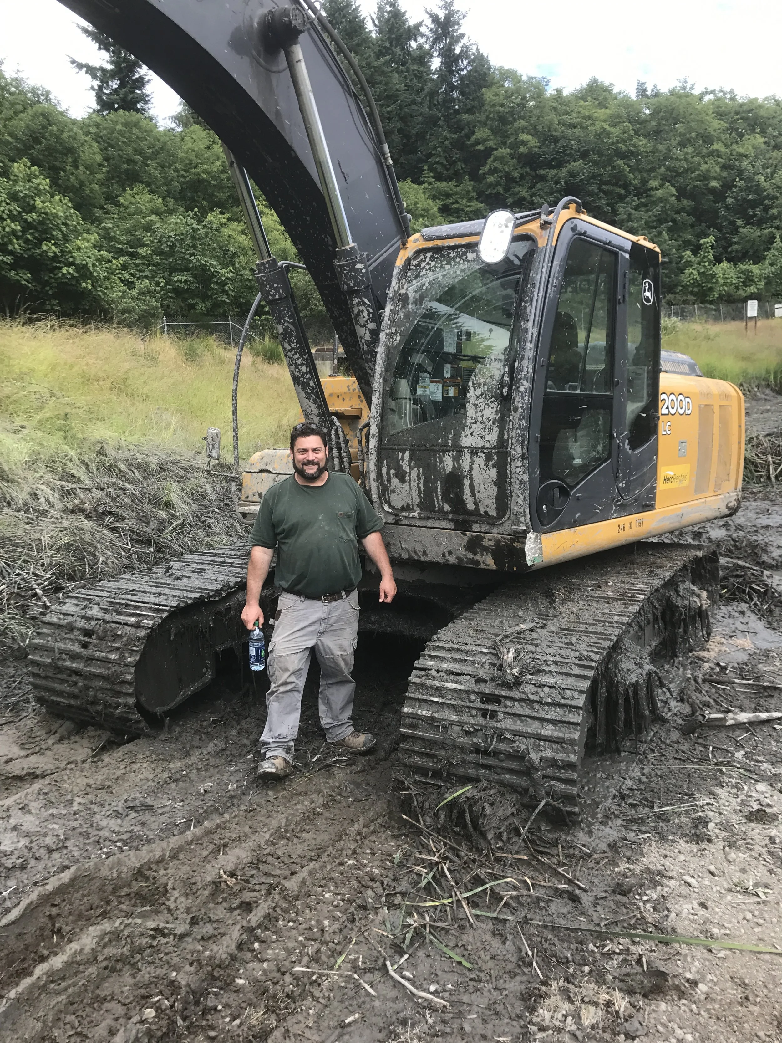 A man standing in front of a yellow excavator at a construction site, with muddy ground and trees in the background.