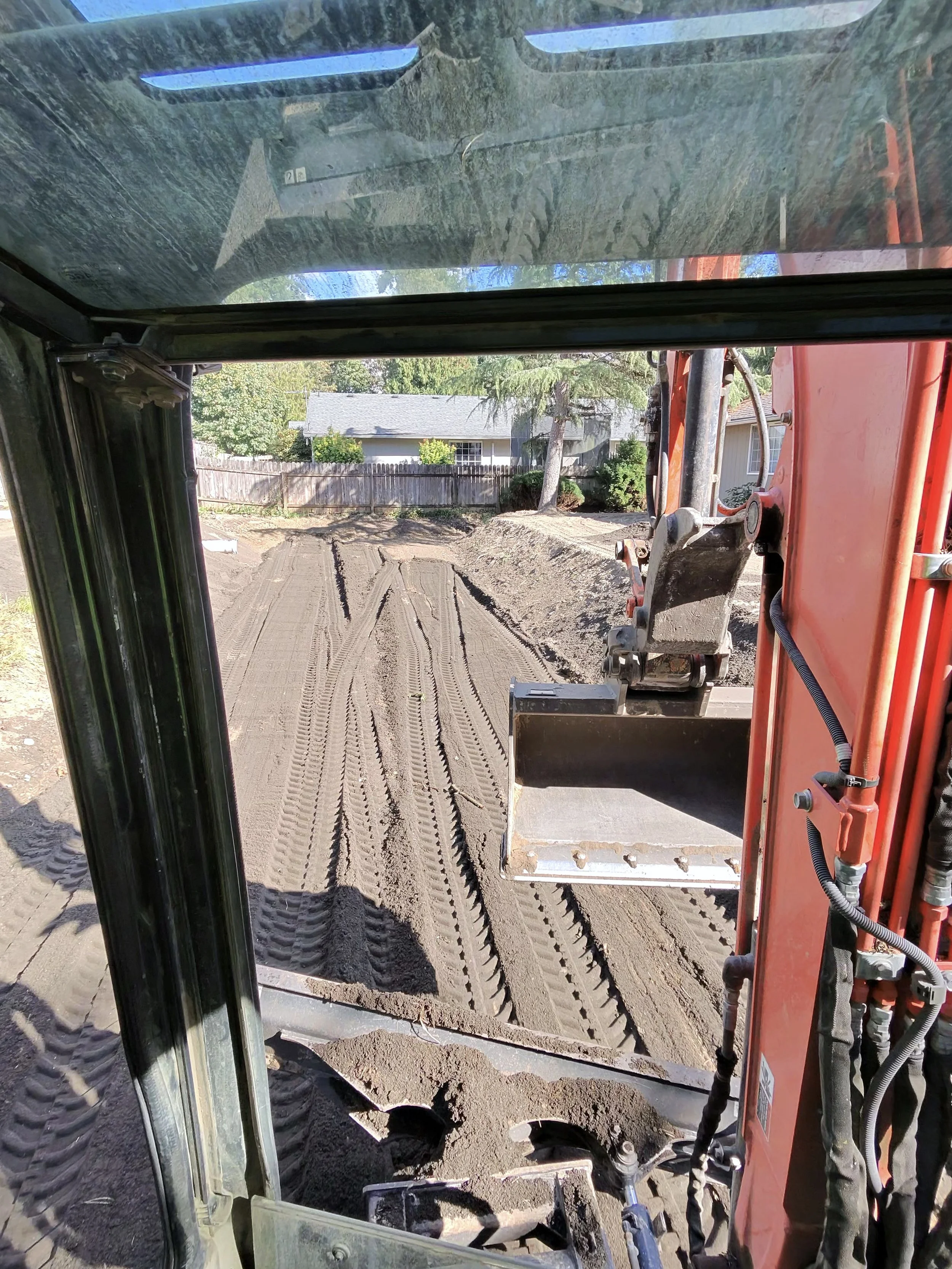 Inside view of a construction vehicle, showing dirt and a construction site with a house and trees in the background, and a shovel bucket attachment.