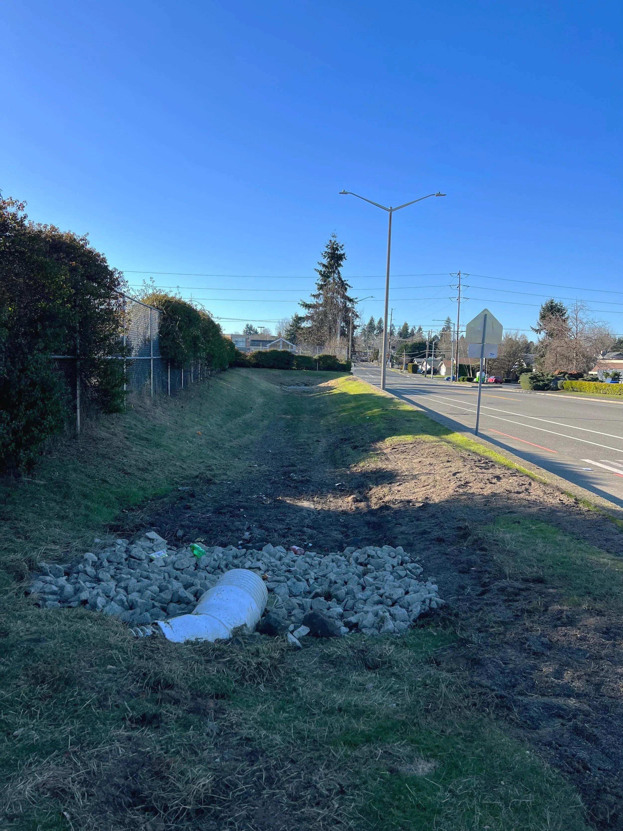 A sidewalk and renovated bioswale with a fence alongside it, and a street with traffic signs, trees, and houses in the background, under a clear blue sky.