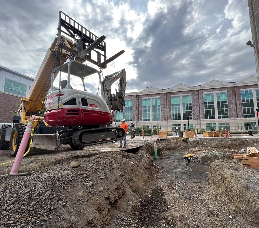Construction site with an excavator and workers near a large building, with a deep trench in the ground.