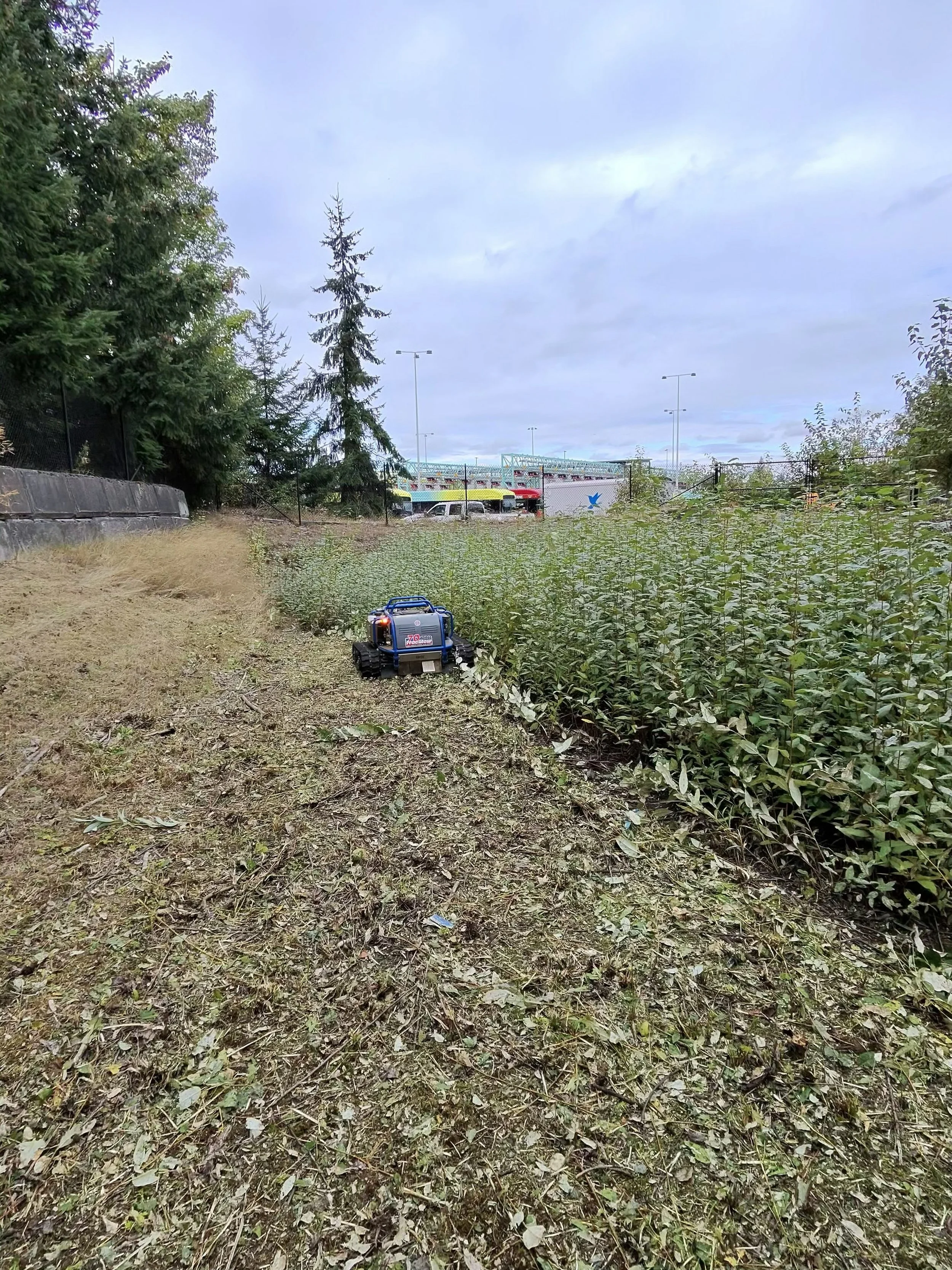 A remote slope mower on the ground in an outdoor area with grass, dirt, and some trees, near a chain-link fence with a parking lot in the background.