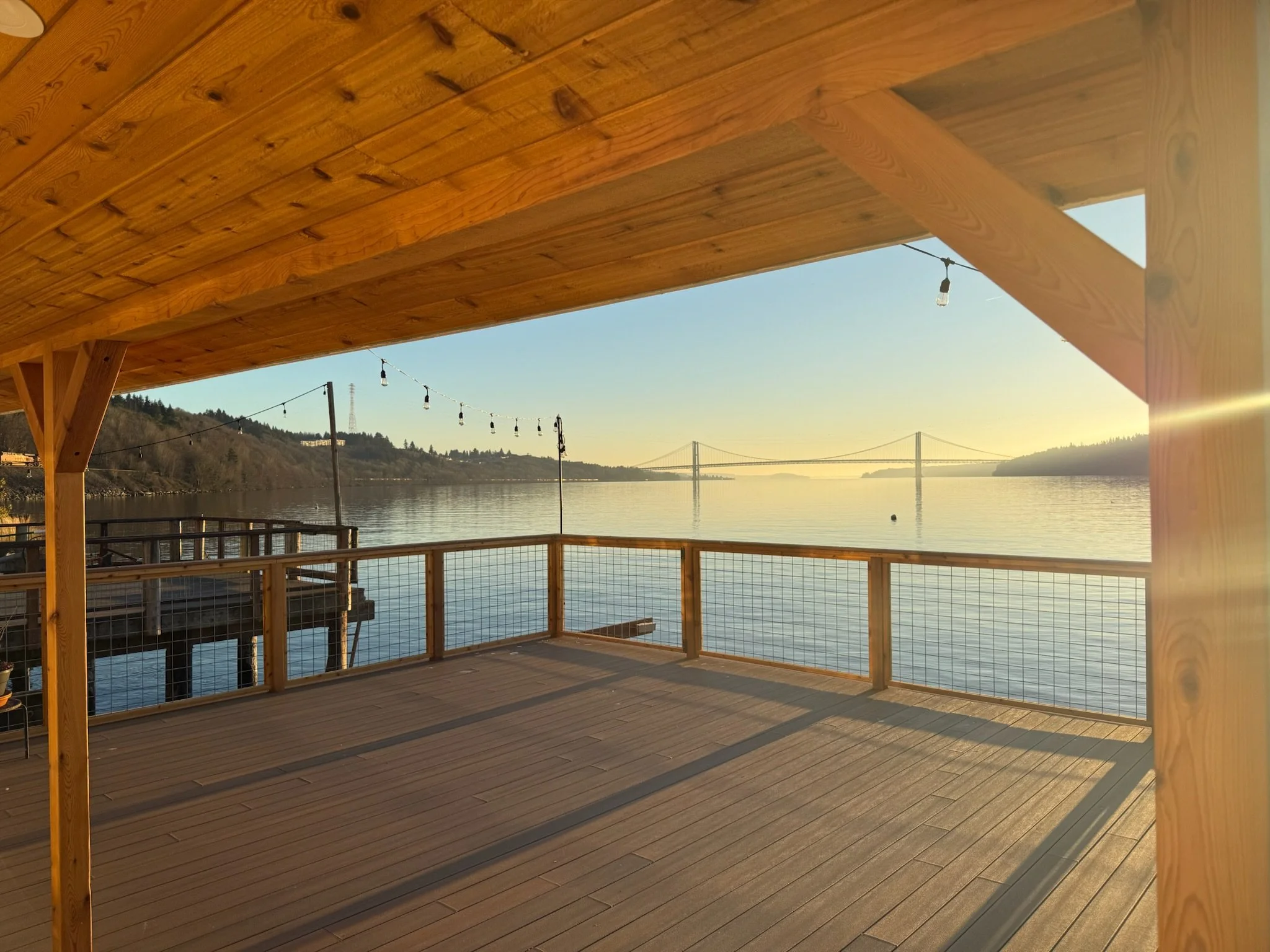 View of a wooden deck overlooking a body of water with hills and a bridge in the distance, under a clear sky during sunset or sunrise.