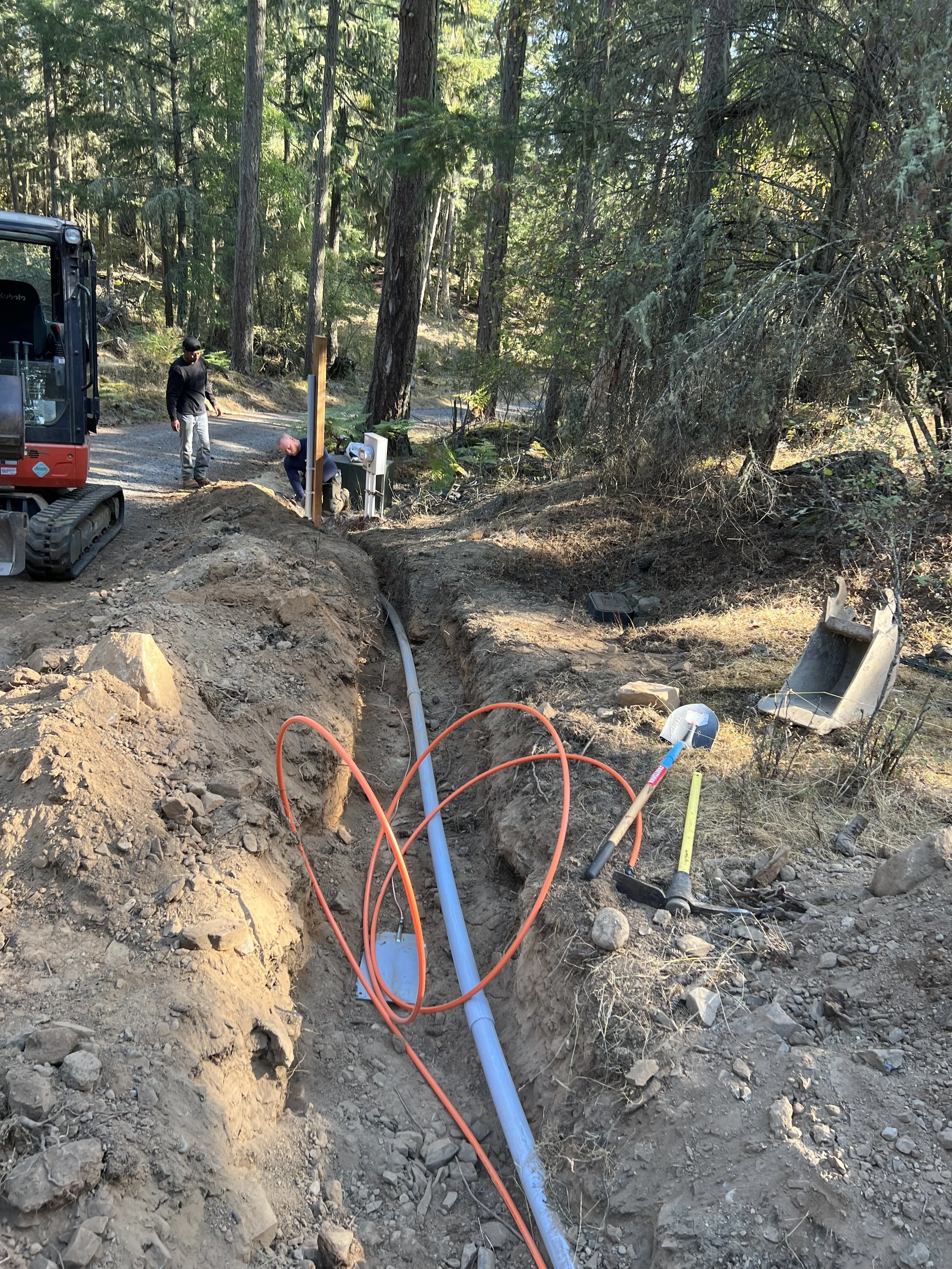 Workers installing underground utility pipes in a forested area with trees and dirt, using tools and equipment.