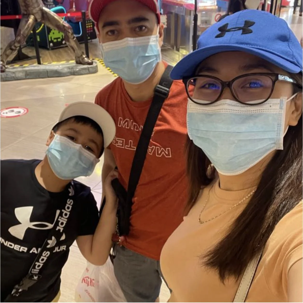 A woman, man, and young boy wearing face masks taking a selfie in a public place. The woman is wearing glasses, a blue cap, and a peach shirt. The man is in a red shirt and red cap, and the boy is in a black Adidas shirt and white cap. There are store displays and a bronze sculpture of a running figure in the background.