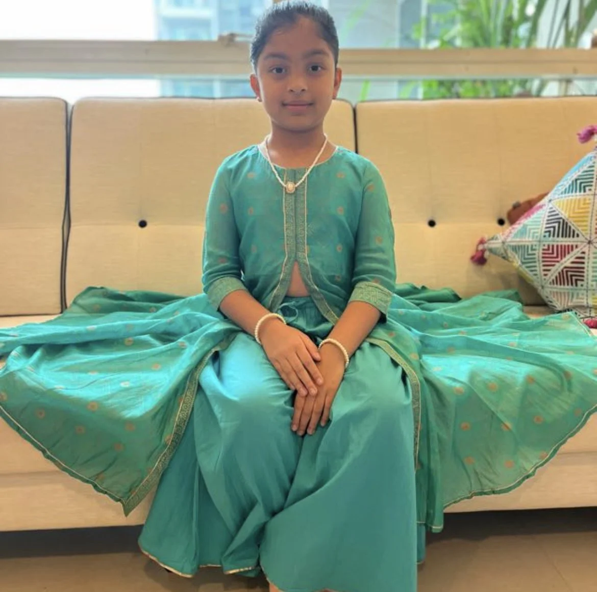 A young girl in a turquoise traditional Indian dress sitting on a beige sofa with a colorful pillow beside her.