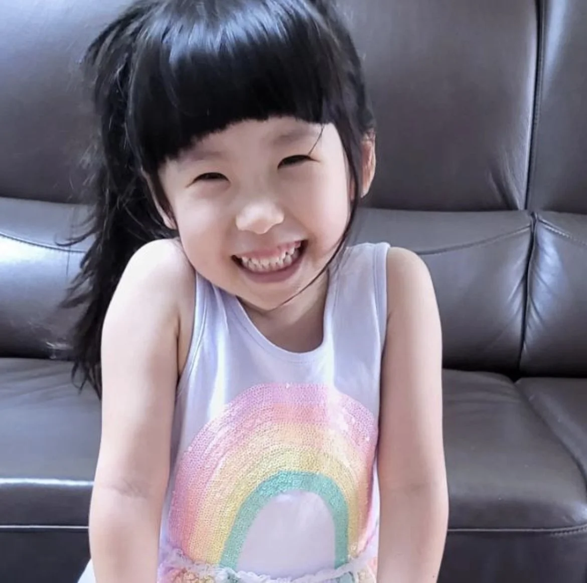 A young girl with black hair smiling widely, showing teeth, wearing a white sleeveless top with a rainbow graphic, sitting on a black leather couch.