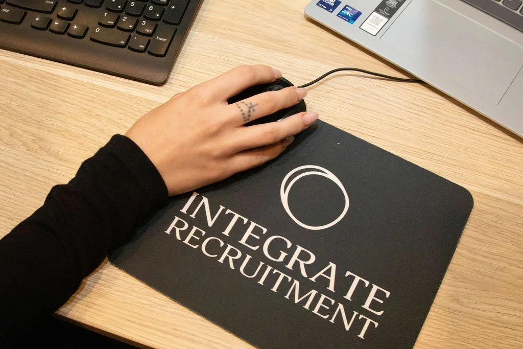 A person's hand is resting on a computer mouse on a mouse pad that reads 'INTEGRATE RECRUITMENT' with a circular logo, on a wooden desk near a keyboard and a laptop.