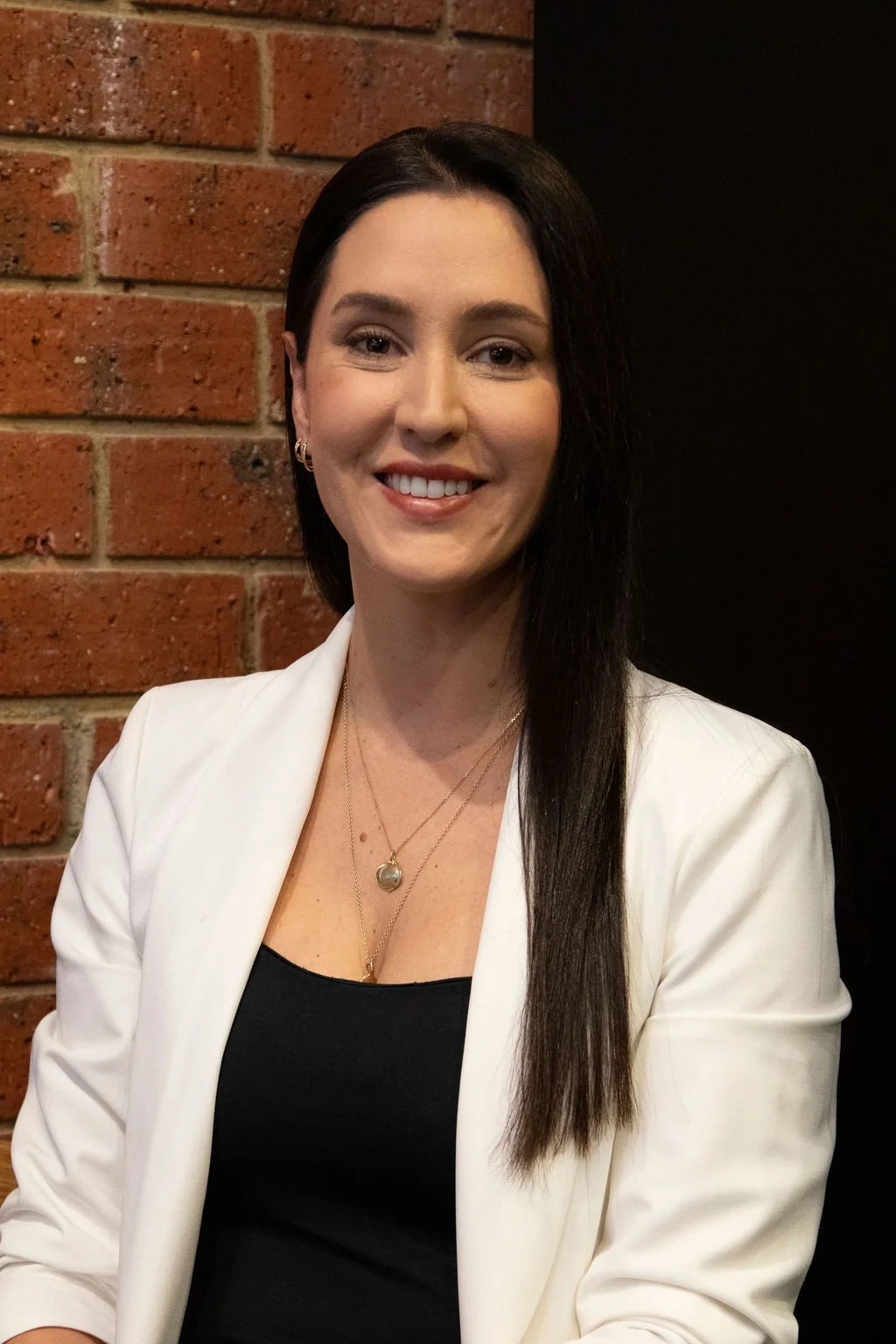 Samantha Bainbridge, smiling, wearing a white blazer over a black top, and layered necklaces, standing against a brick wall.