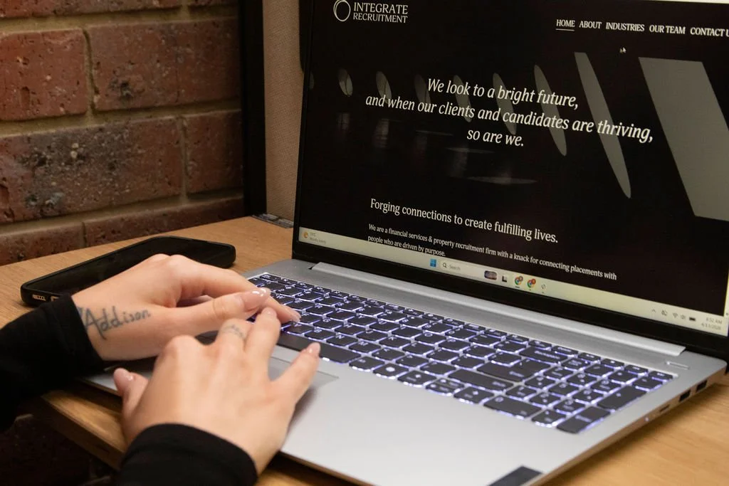 Person typing on a laptop keyboard in front of a brick wall, with a smartphone on the wooden desk and a website related to recruitment displayed on the screen.