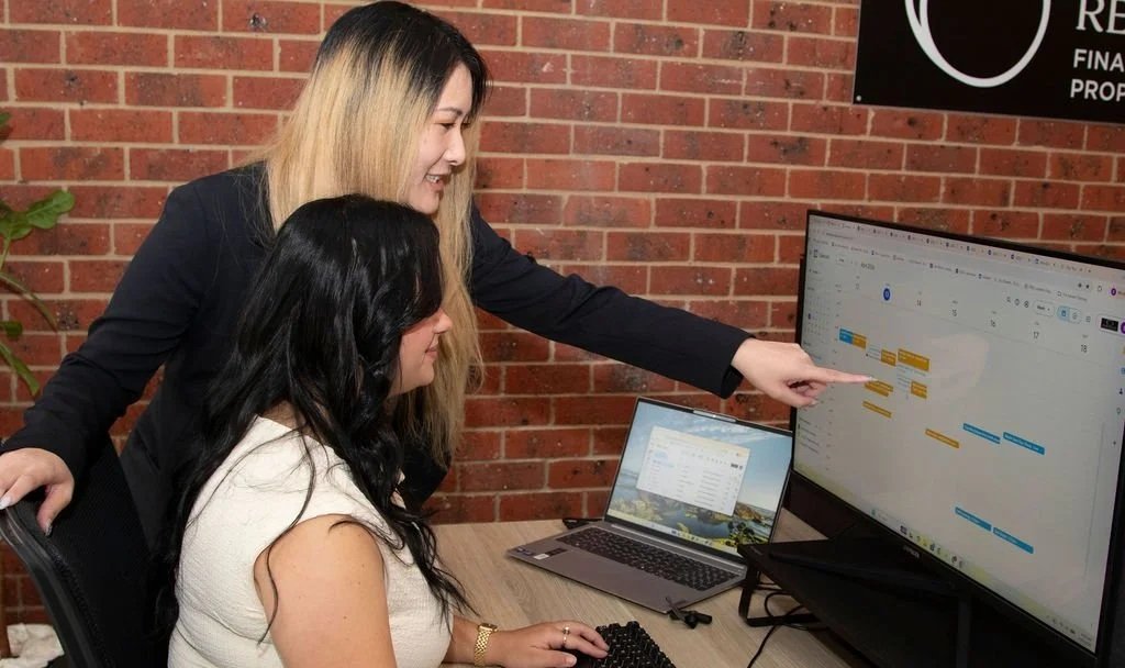 Two Integrate Recruitment Staff members working together on a project, with one pointing at a computer screen displaying a gantt chart or project timeline.
