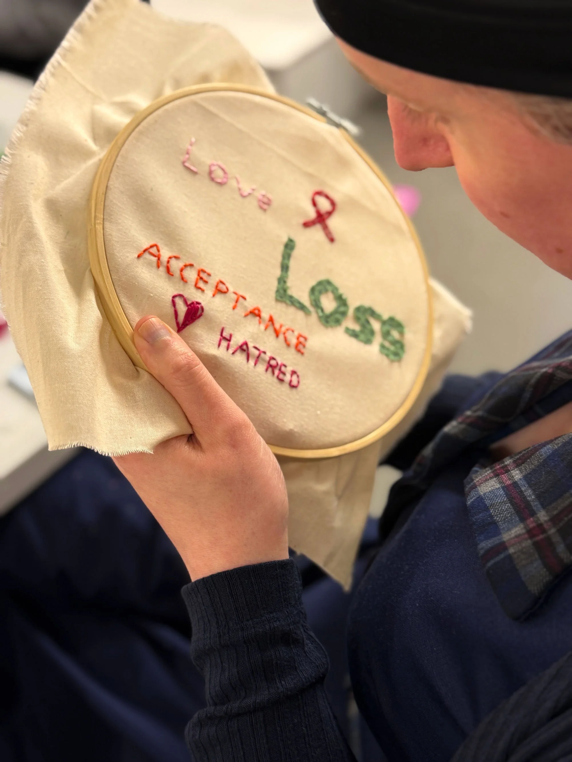 Participant stitching the words “Love & Loss” alongside “Acceptance” and “Hatred” in an embroidery hoop.