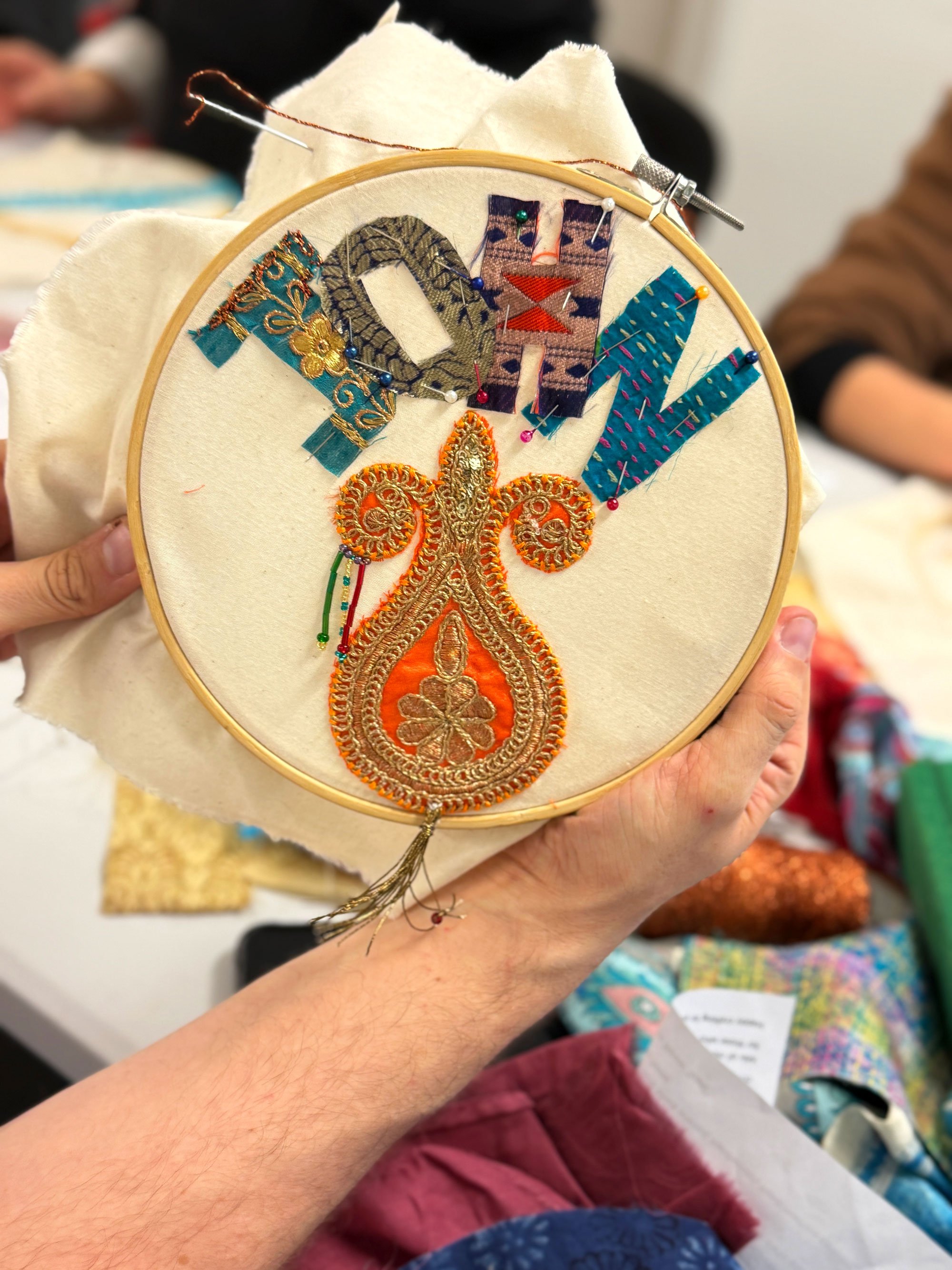Embroidery hoop with layered textile letters and ornate gold appliqué reading “JOHN.”