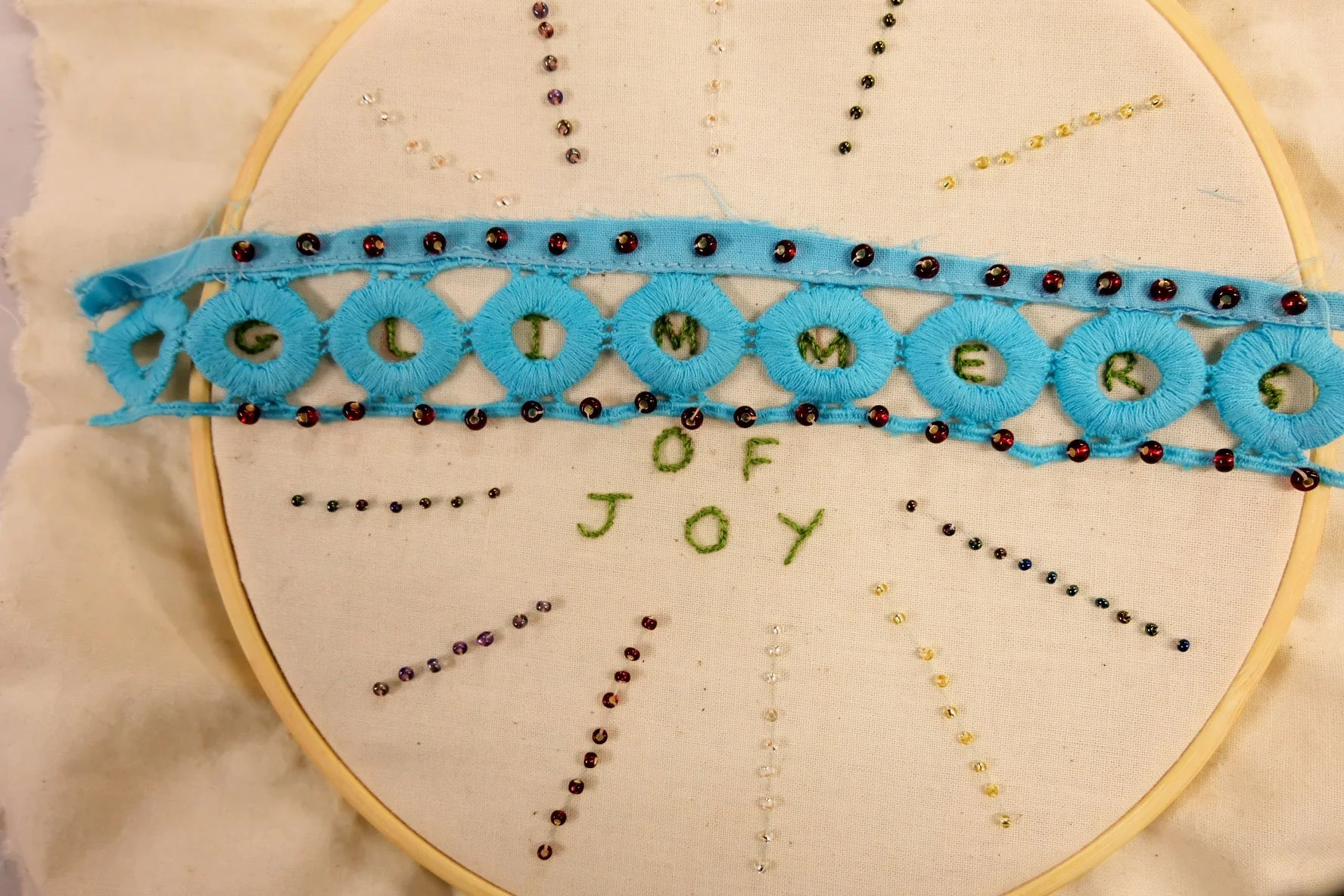 Embroidery hoop featuring stitched text “Glimmers of Joy” surrounded by beads and blue lace trim.
