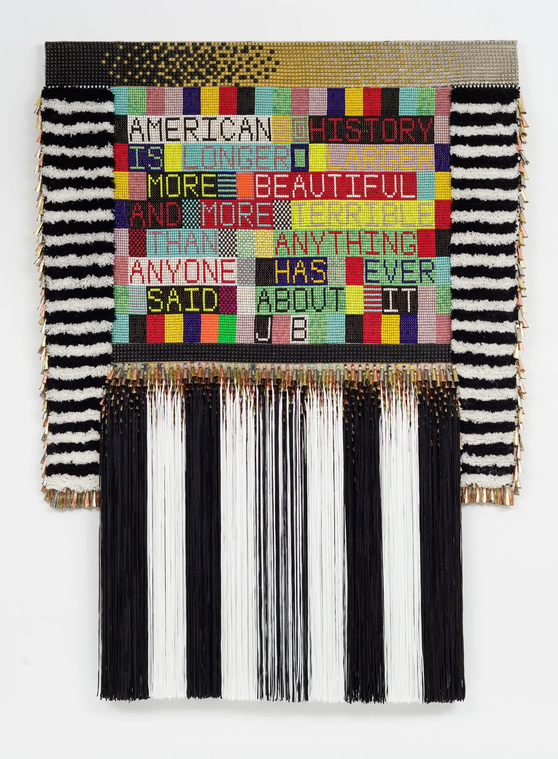 Large textile wall hanging by Jeffrey Gibson featuring bold beaded text and long black and white fringe.